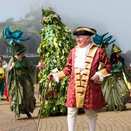 Man dressed as a historical figure in a red and gold coat, with women dressed as mythical plants or trees holding large blue flower props, standing outdoors.