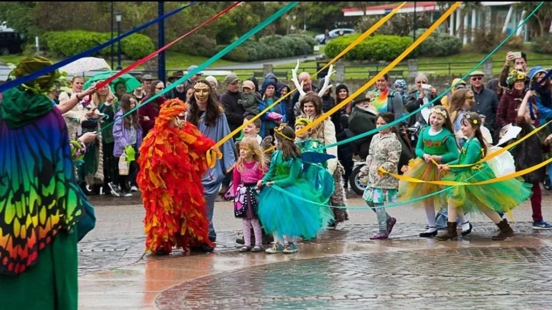 Festive outdoor event with children and adults, some in colorful costumes and fairy wings, playing with ribbons on a wet brick surface, with onlookers in the background.