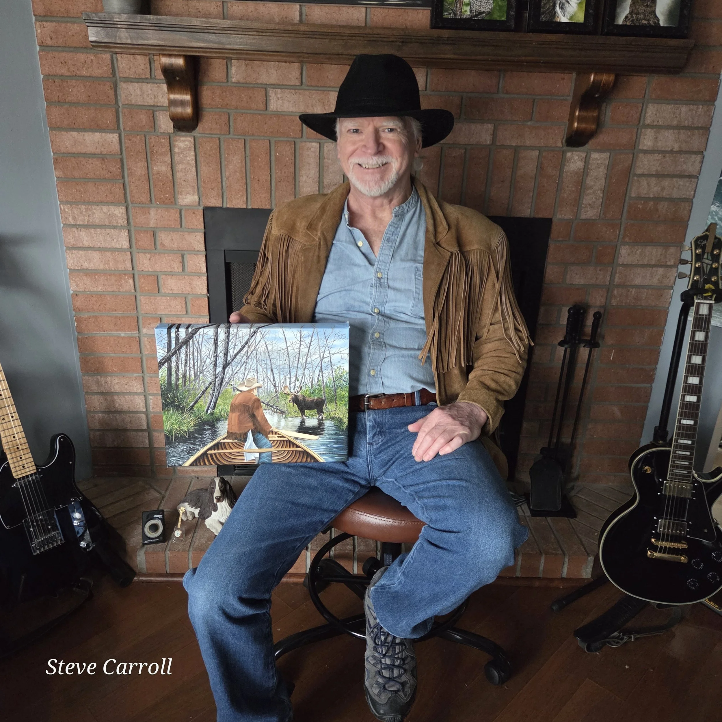 Steve Carroll sitting on a brown chair in his den while holding an oil painting of himself in a canoe on a remote river in Ontario. The river is blocked by a fallen dead tree as well as a moose standing in the river.