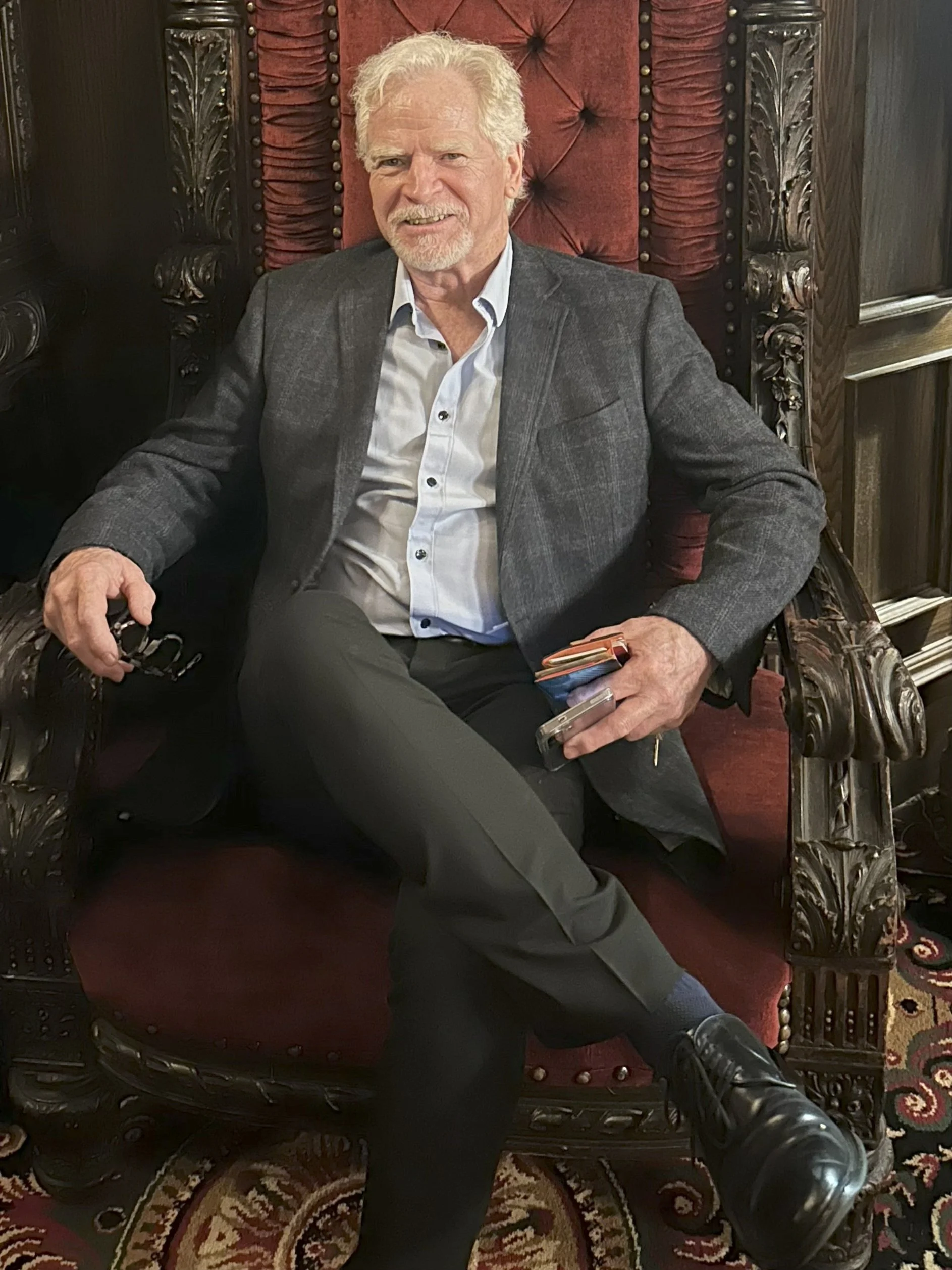 Artist Steve Carroll wearing a gray blazer and light shirt, sitting on a large, ornate wooden chair with a red cushion, in a richly decorated room after a successful art show.
