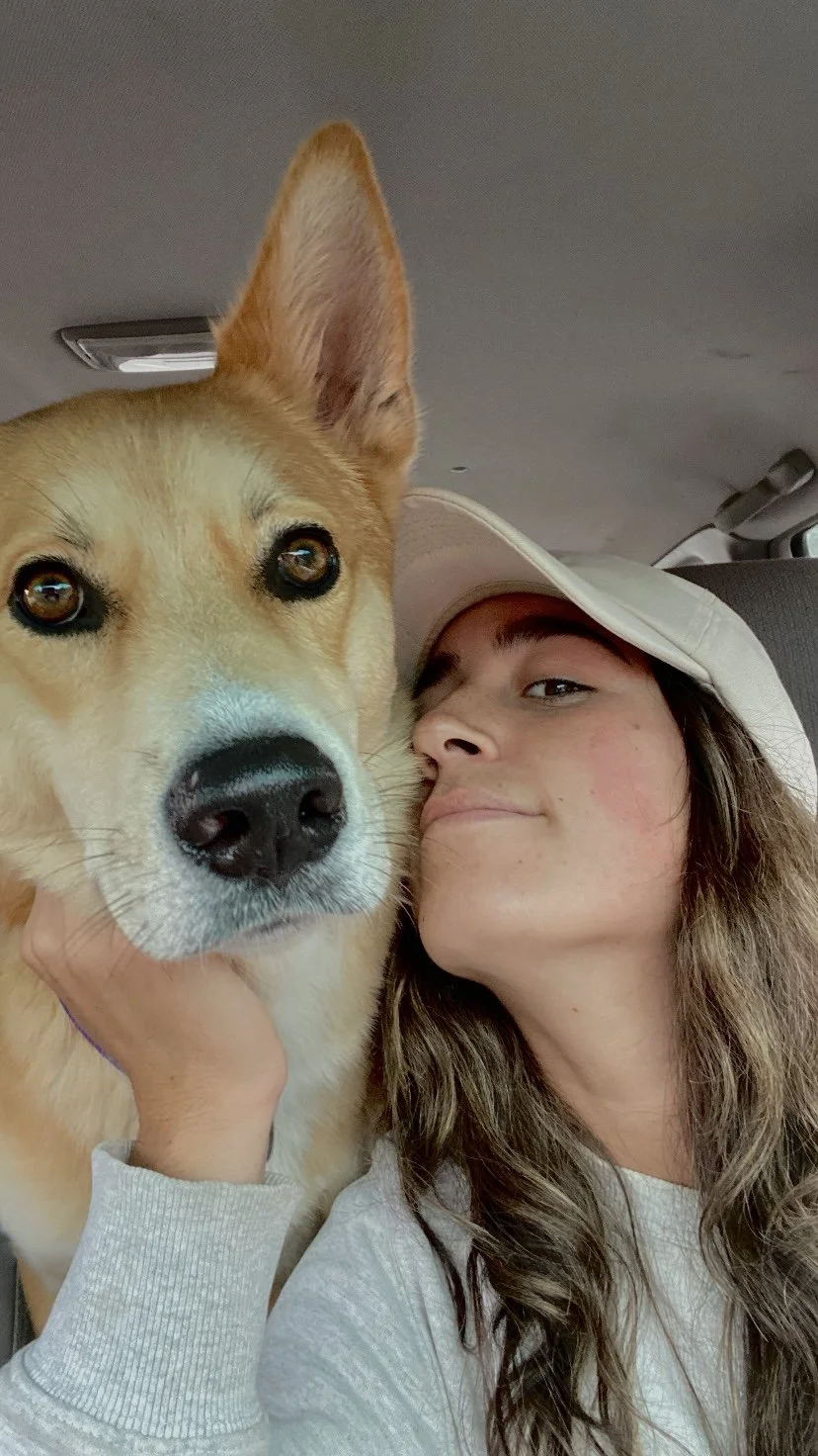 A woman with long wavy hair and a beige baseball cap taking a selfie with her dog, a Golden Retriever mix, inside a vehicle.
