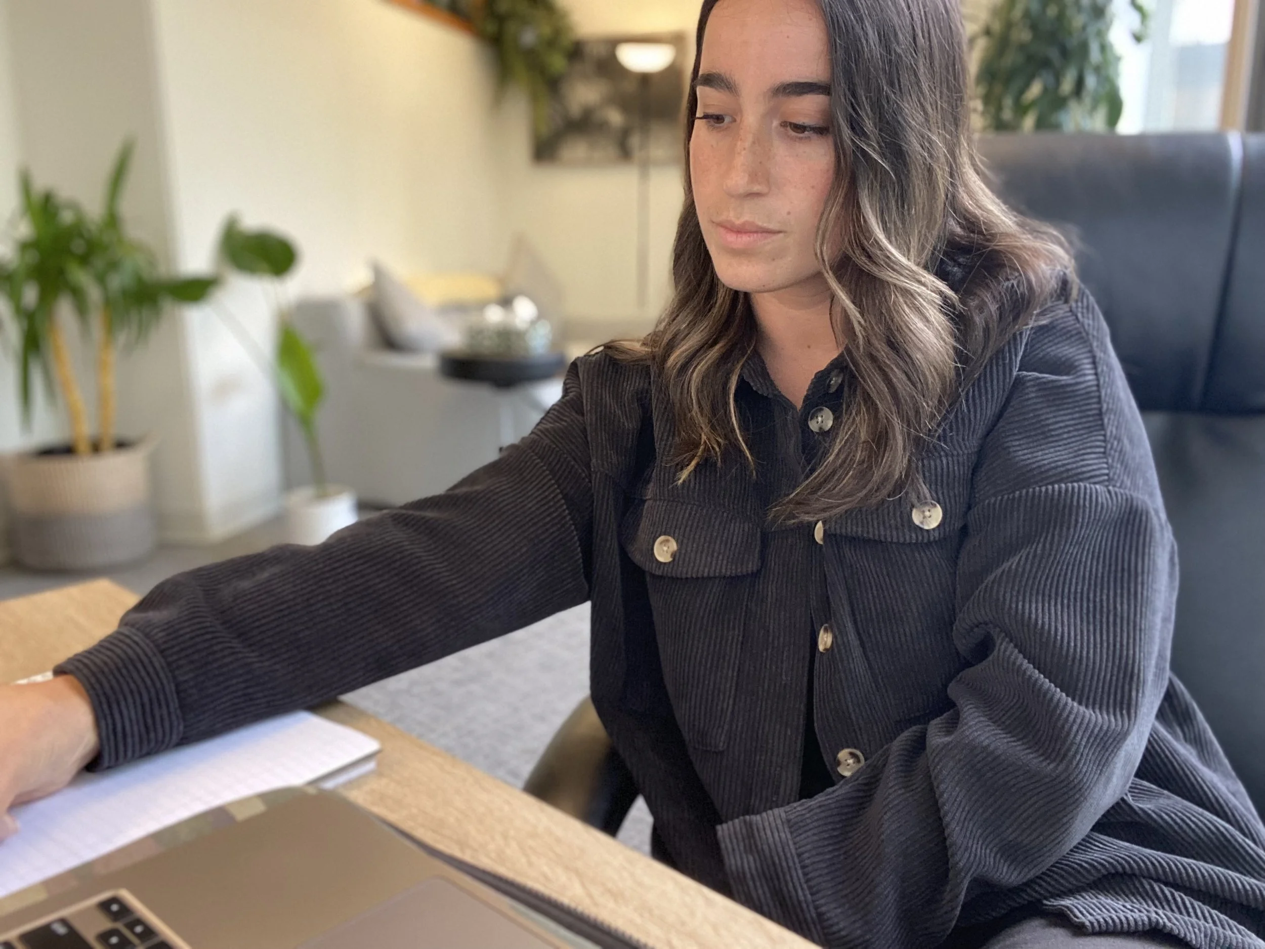 A woman with wavy brown hair working at a desk with a laptop and notebook, in a modern office with plants and artwork in the background.