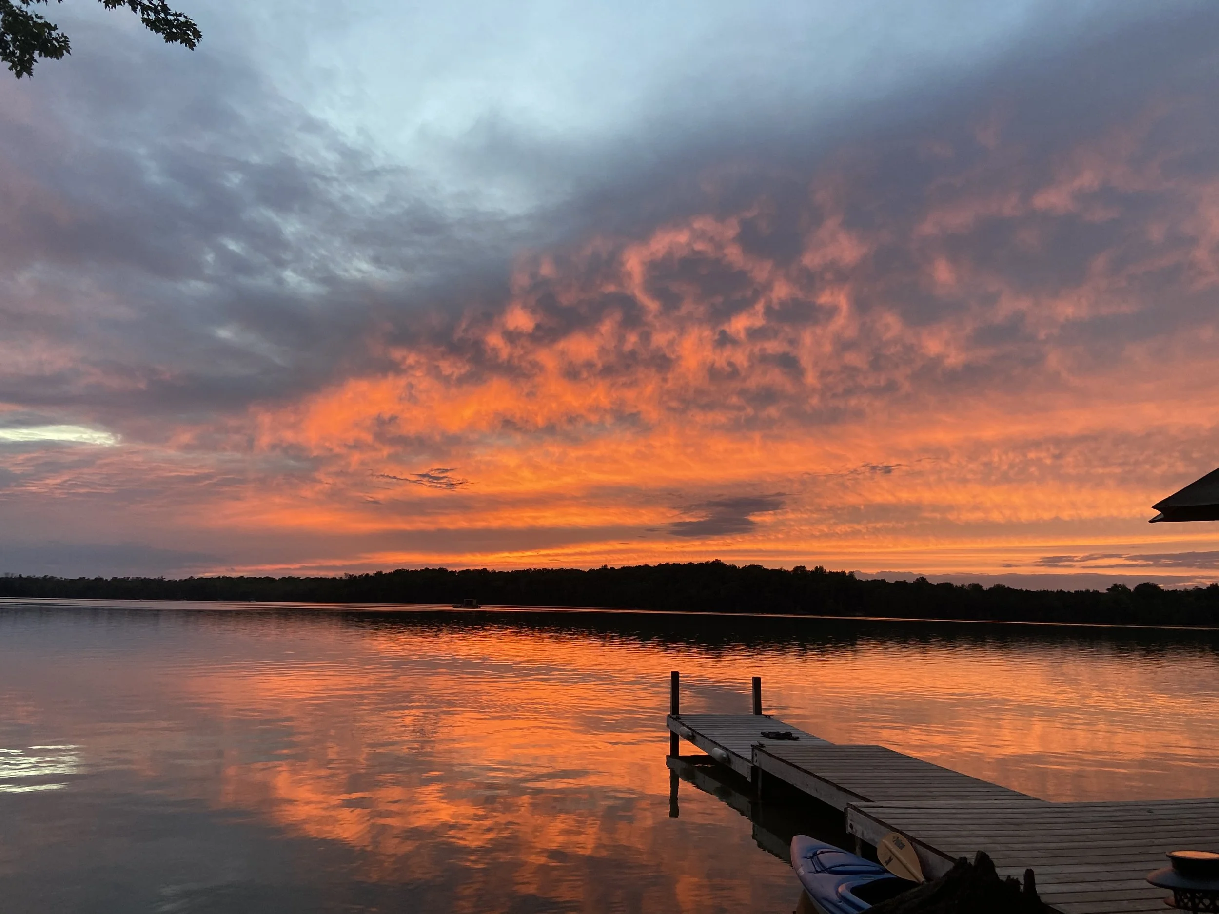 Sunset over a calm lake with vibrant orange and pink clouds, a pier extending into the water, and a kayak on the shore.
