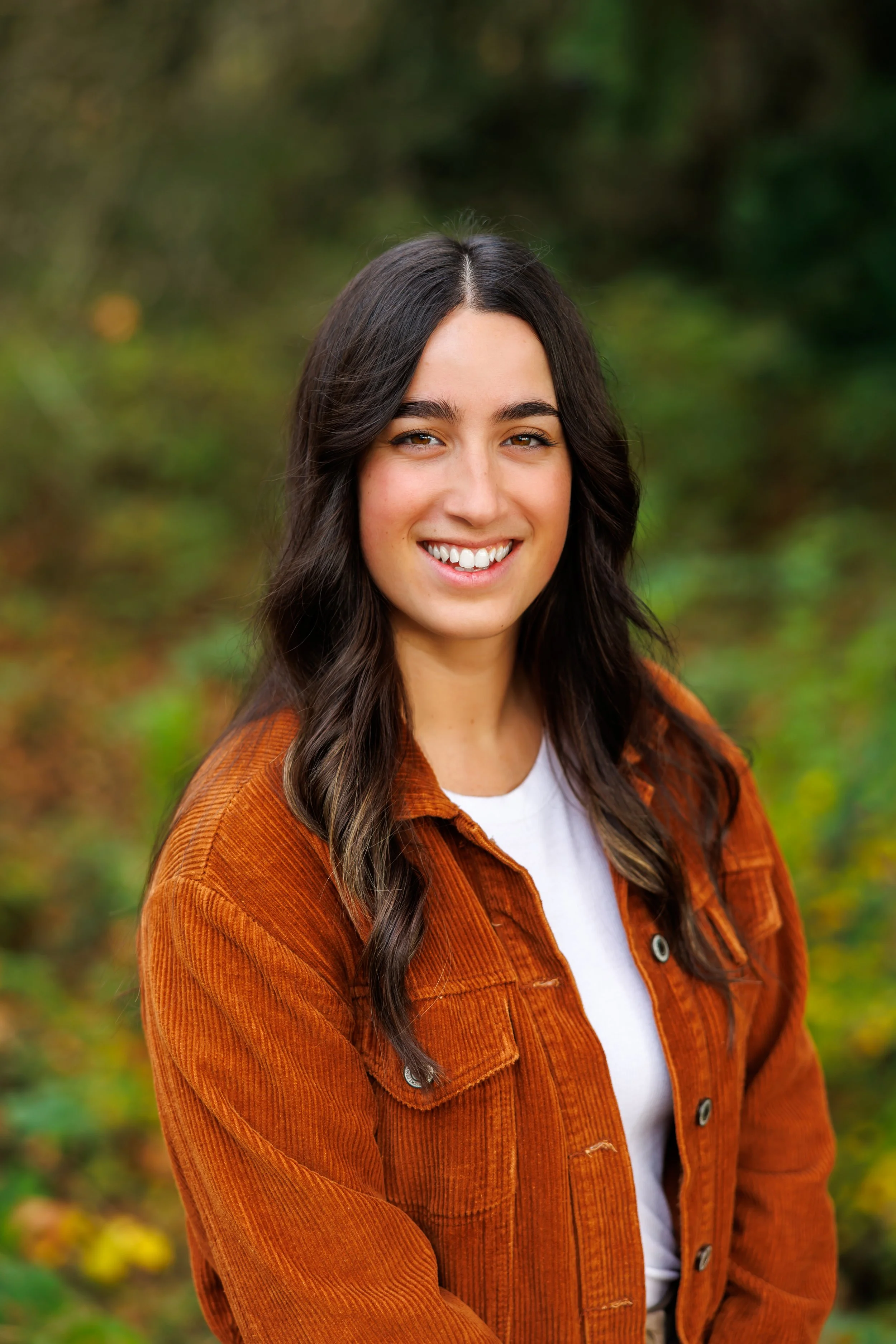 A young woman with long dark hair, smiling, wearing a rust-colored corduroy jacket and a white shirt, standing outdoors with blurred green foliage in the background.