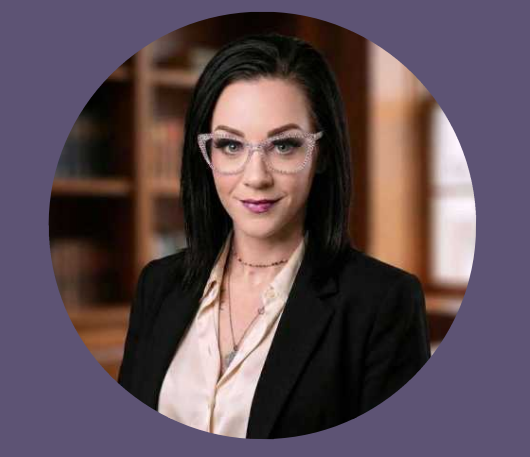 A woman with straight black hair wearing glasses, a black blazer, and a white blouse, standing in a room with wooden shelves in the background.