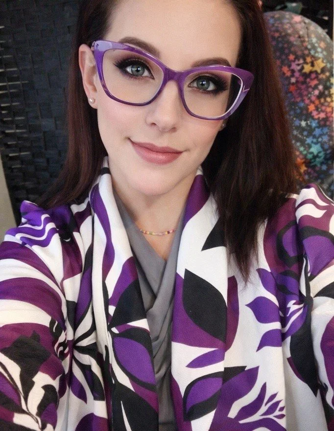 A woman wearing large purple glasses, a colorful patterned jacket with purple, black, and white colors, and a grey top, smiling at the camera.