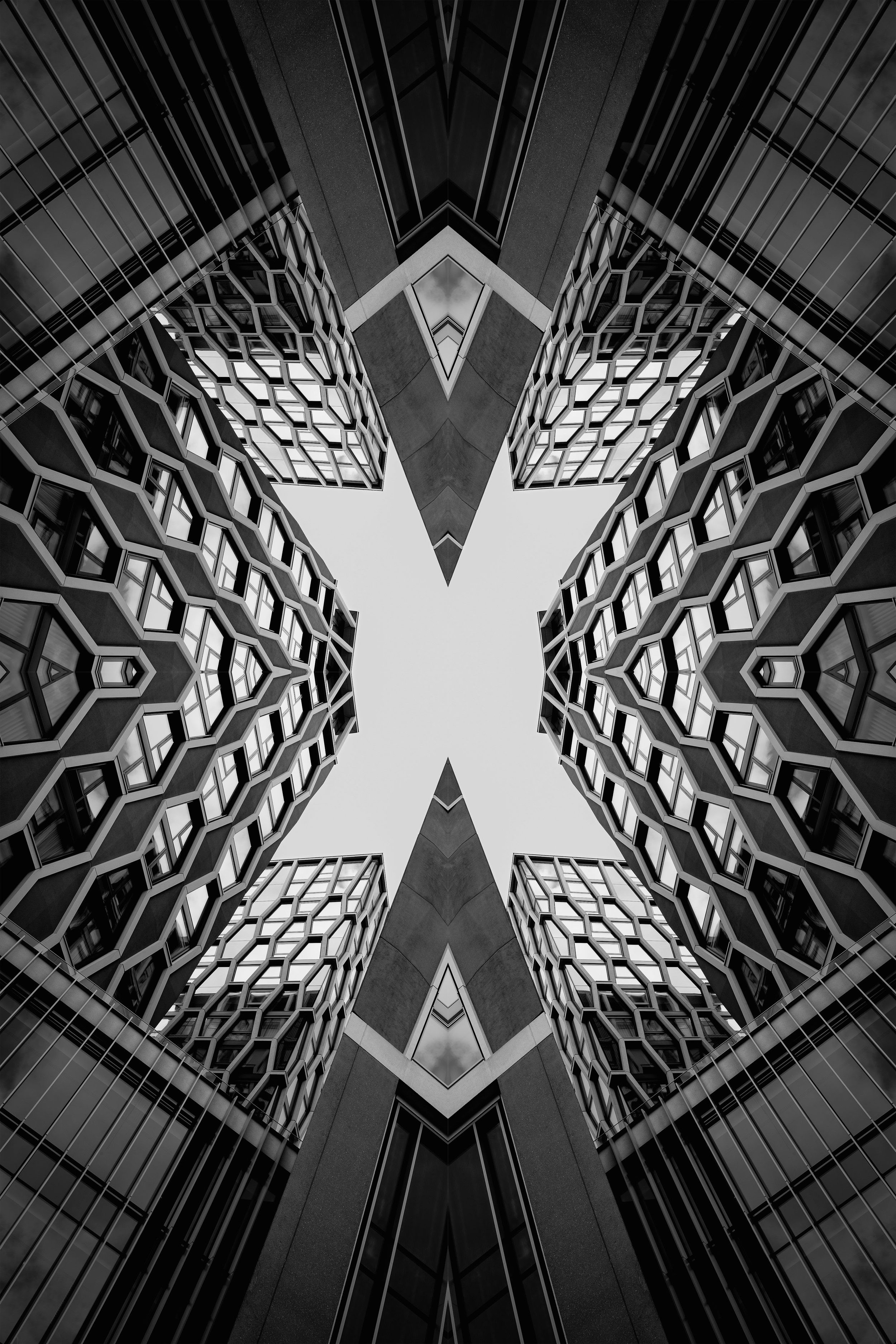 A black and white photo of tall modern buildings with geometric windows, taken from the ground looking upward, creating a symmetrical pattern with a view of the sky in the center.