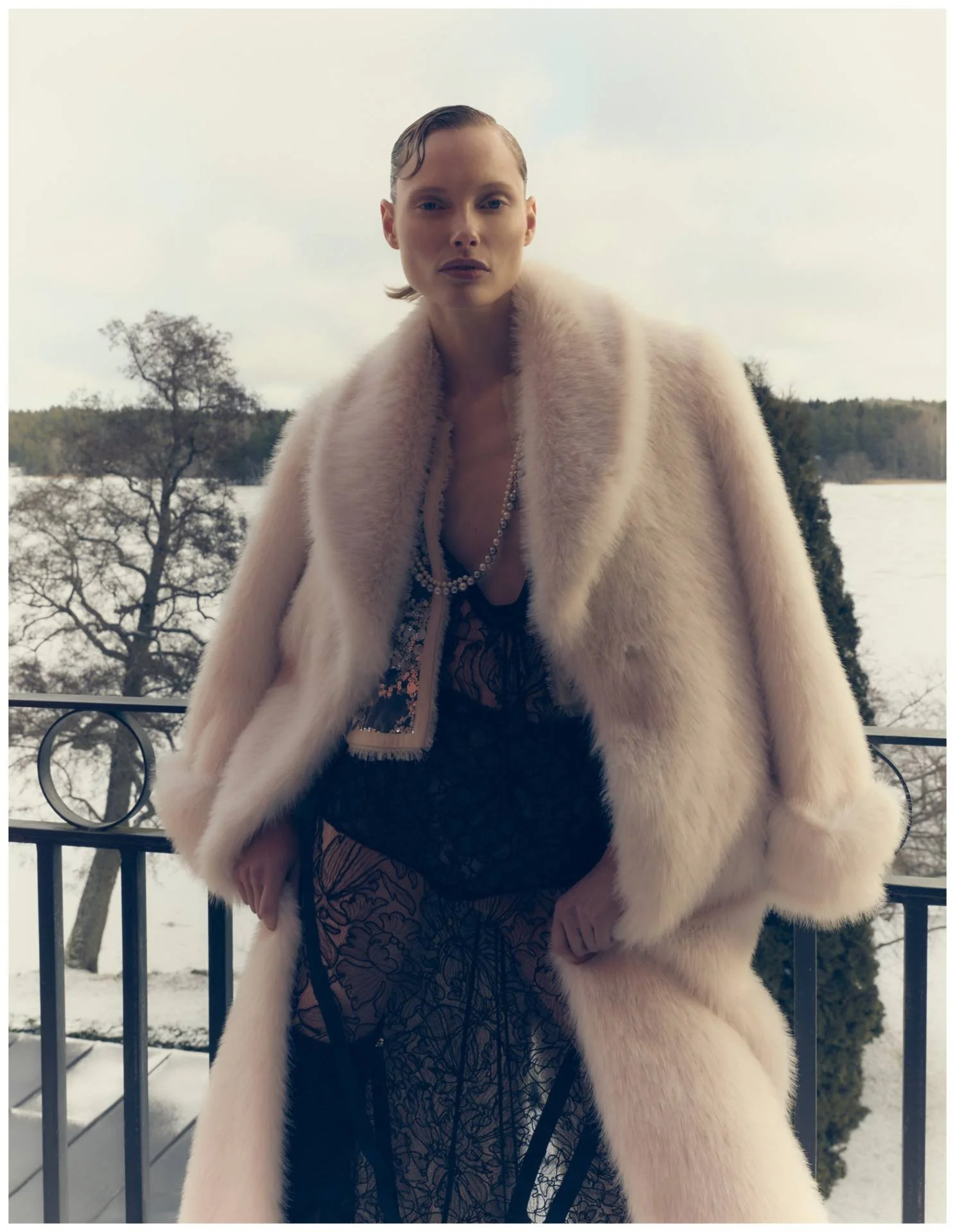 A woman standing outdoors on a balcony wearing a luxurious cream-colored fur coat, black lace dress, and layered pearl necklaces with a snow-covered landscape and trees in the background.