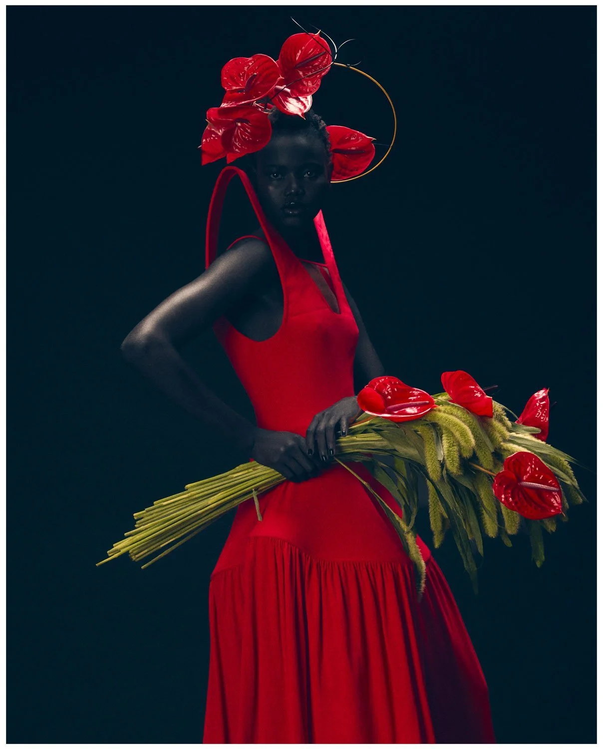 A woman dressed in a red dress holding a bouquet of red anthuriums and greenery, with a large flower headpiece and a gold hoop accessory, against a dark background.