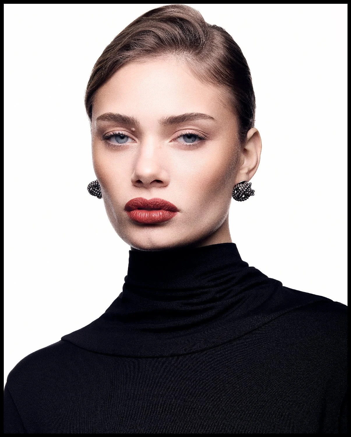 Portrait of a woman with short brown hair, wearing a black turtleneck and black earrings, against a white background.