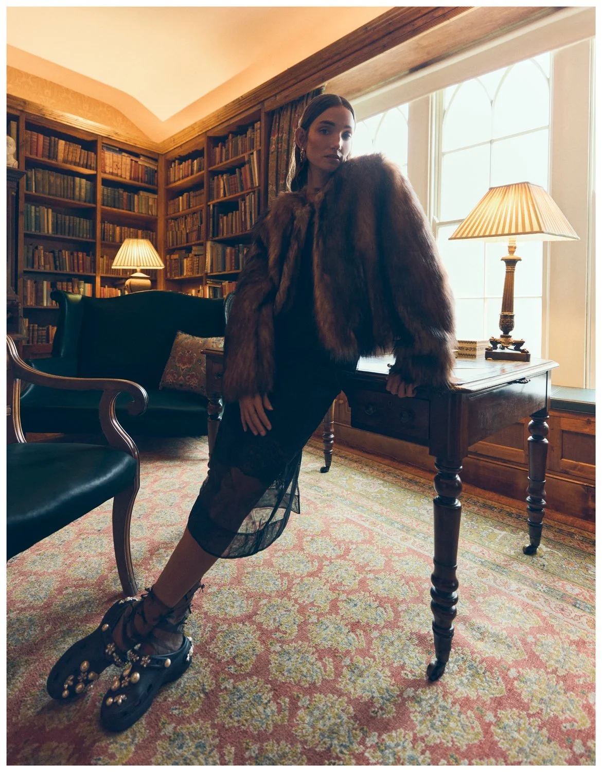 A woman posing in a vintage-style library with bookshelves, a table with a lamp, and a window with curtains, wearing a fur coat, mesh dress, and decorative shoes.