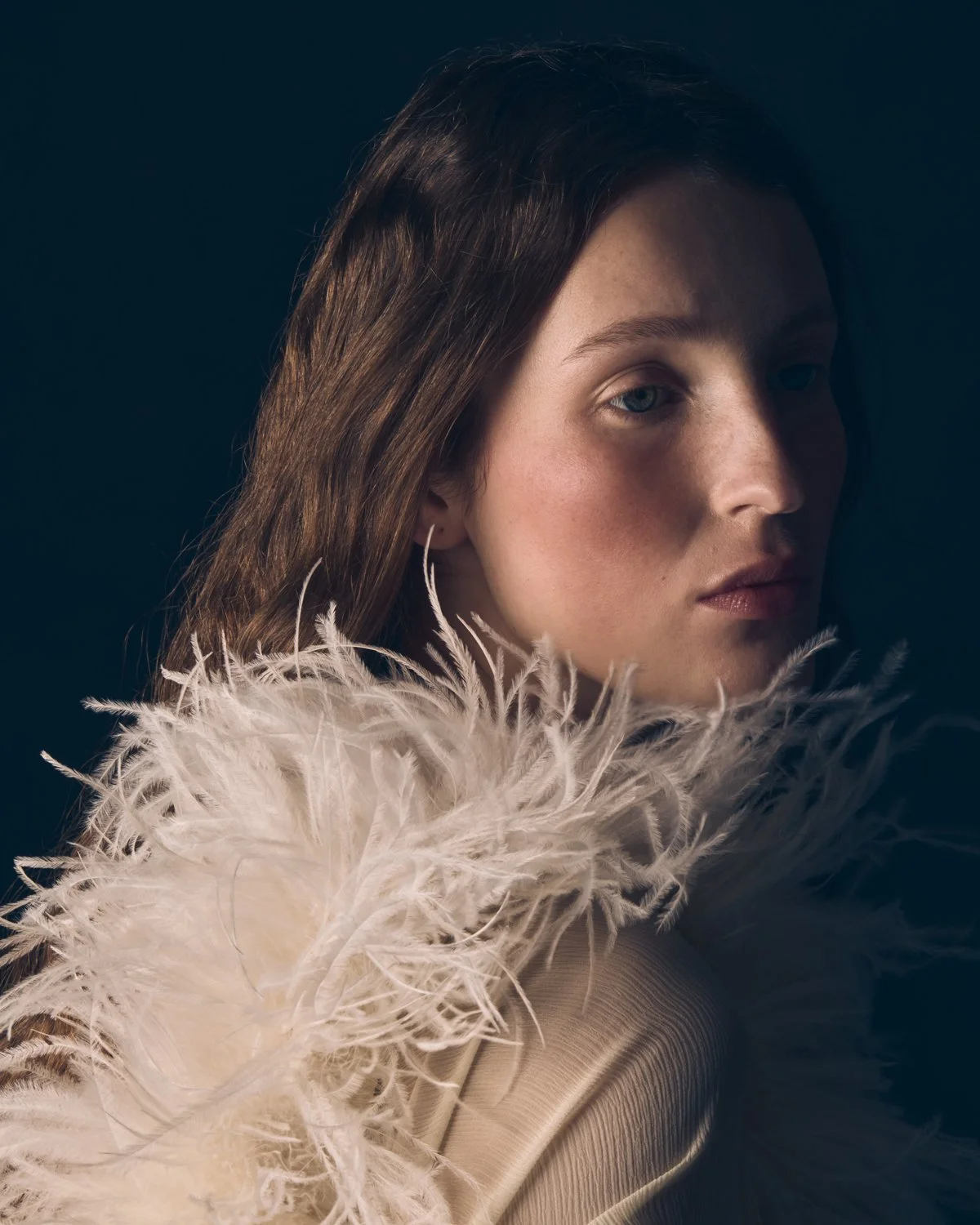 Portrait of a woman with long brown hair wearing a beige dress with feather details, against a dark background.
