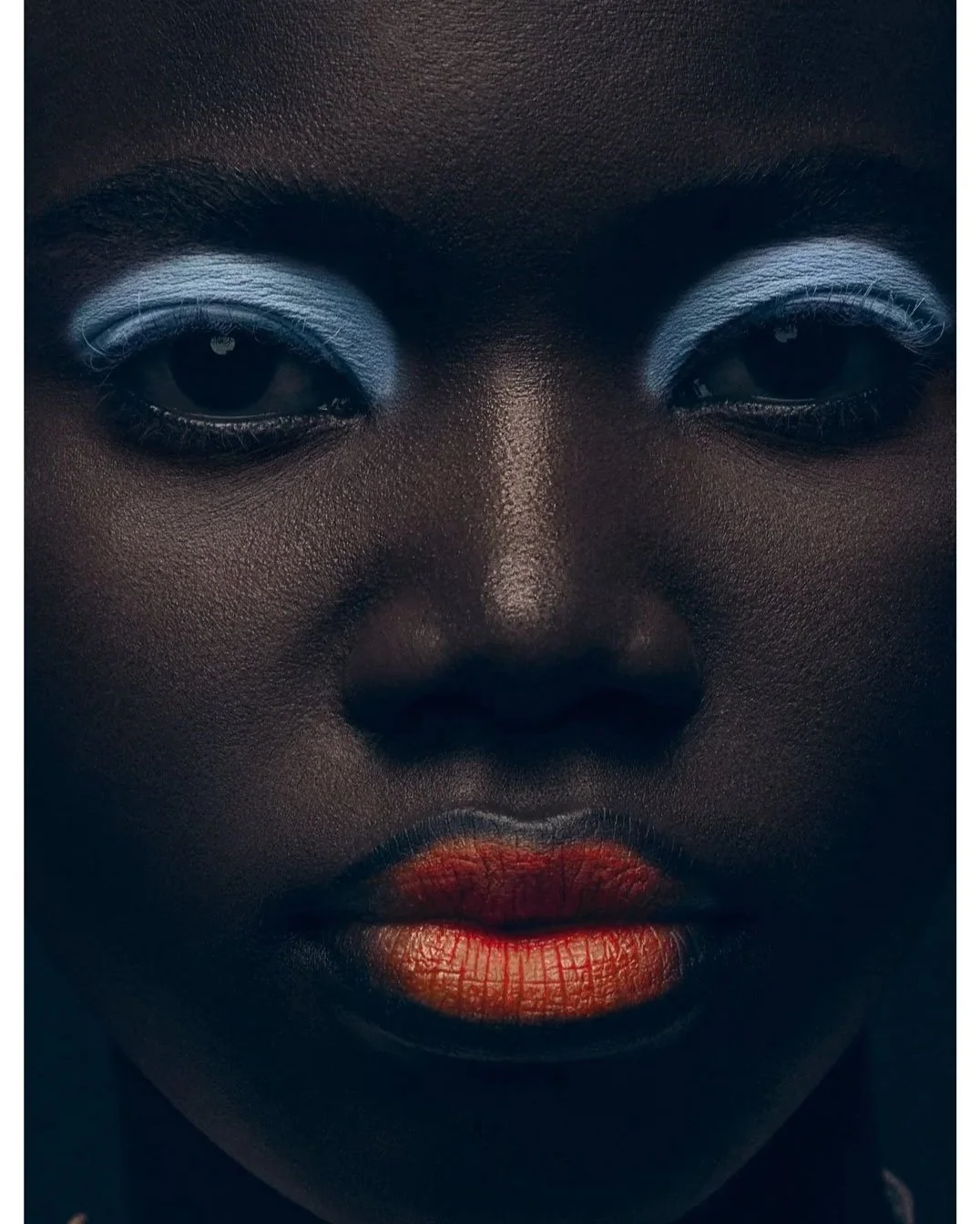 Close-up of a woman's face with dark skin, silver eyeshadow, red lipstick, and dark eyes.