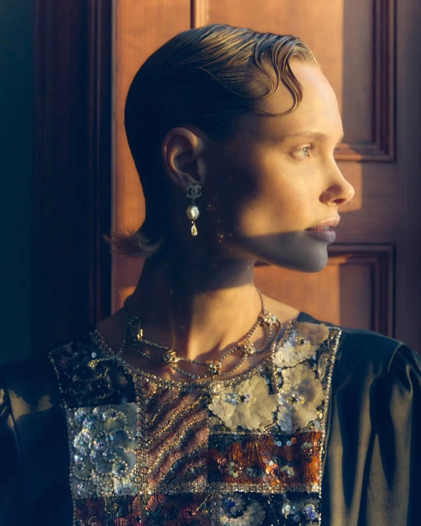 Profile of a woman with short, wavy hair, wearing pearl earrings and layered necklaces, dressed in a jeweled dress, standing by a wooden wall, illuminated by warm sunlight.