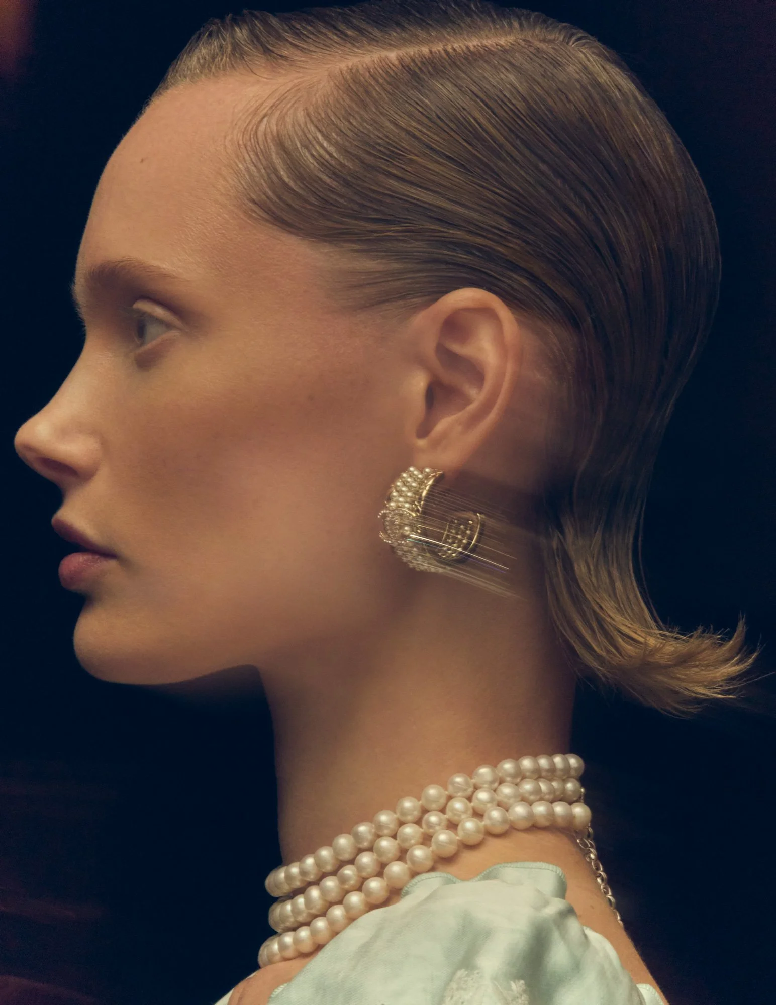 Side profile of a woman with slicked-back hair, wearing pearl earrings, a pearl necklace, and a satin blouse, against a black background.