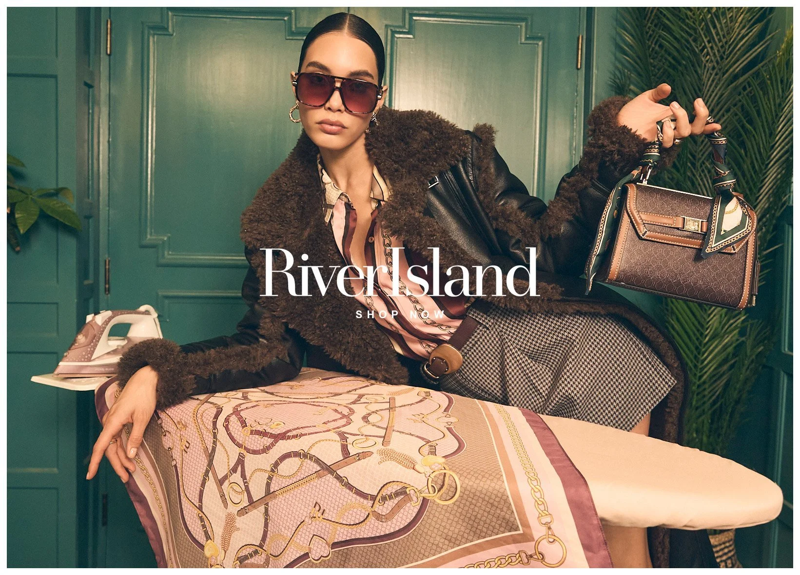 A woman with dark hair, wearing sunglasses, a patterned shirt, and a black leather jacket with fur trim, is sitting at an ironing board covered with a scarf, holding a handbag, with a vintage telephone nearby against a green wall with plants.