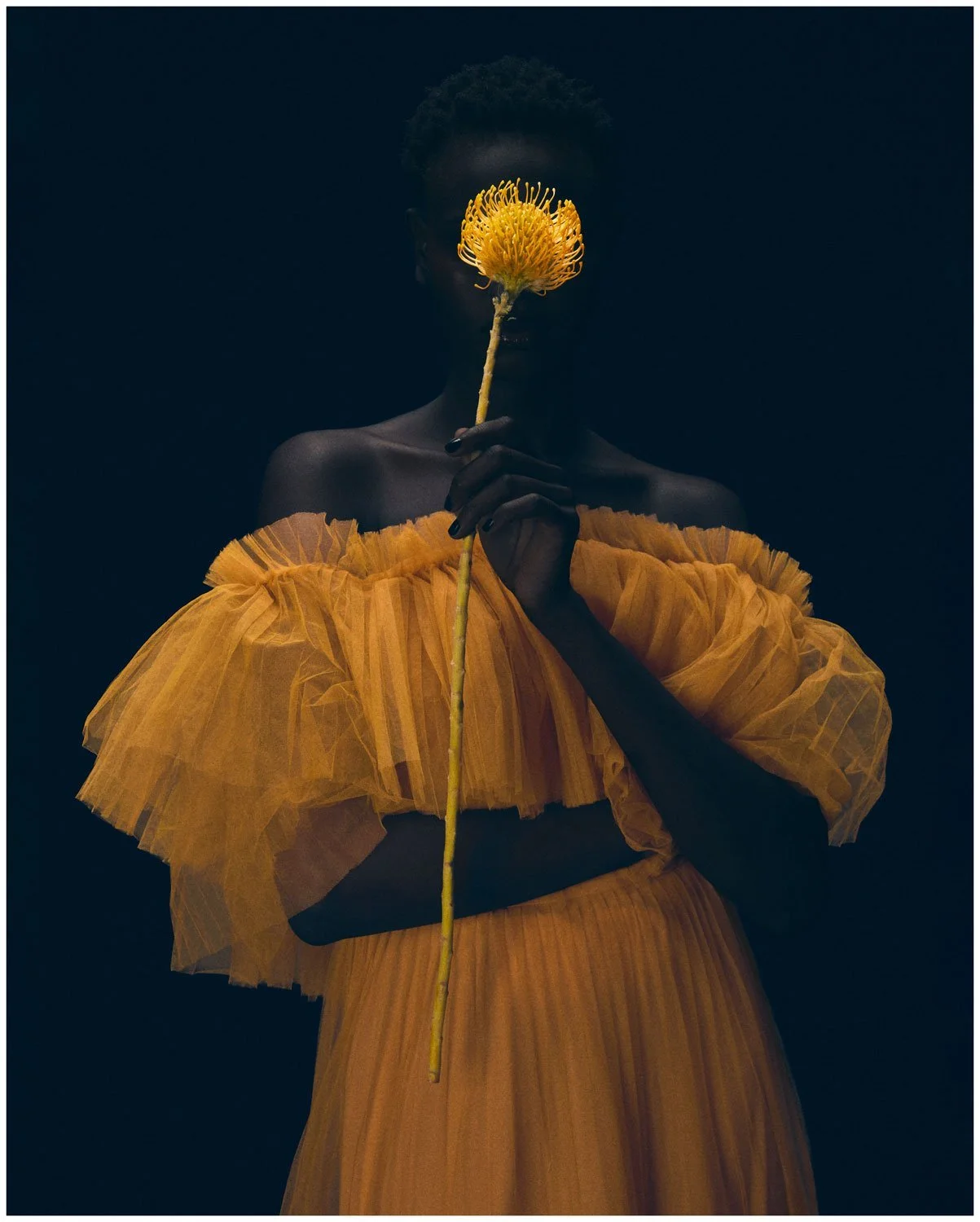 A person dressed in a yellow off-the-shoulder dress holding a yellow flower in front of their face, with their face partially obscured by the flower, against a dark background.