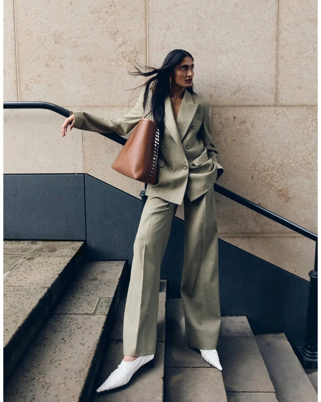 A woman in a beige suit with wide-leg pants is standing on stairs against a beige wall, holding a brown handbag, with her black hair flowing and wearing white pointed shoes.