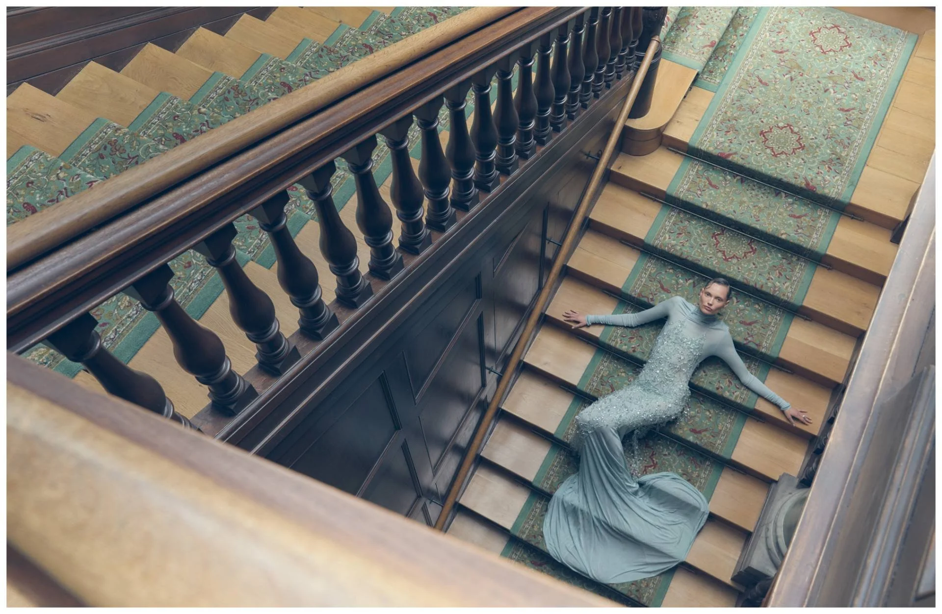 A woman in a sparkly, fitted gown lying on a wooden staircase with ornate carpets, captured from above.