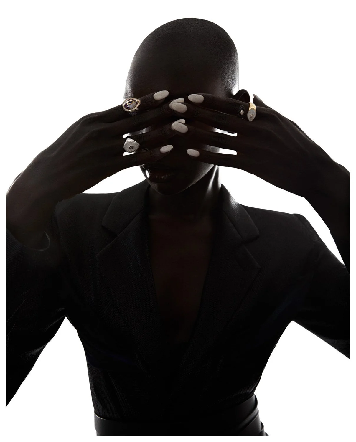 A person with a shaved head covering their eyes with their hands, wearing rings and a dark blazer against a white background.