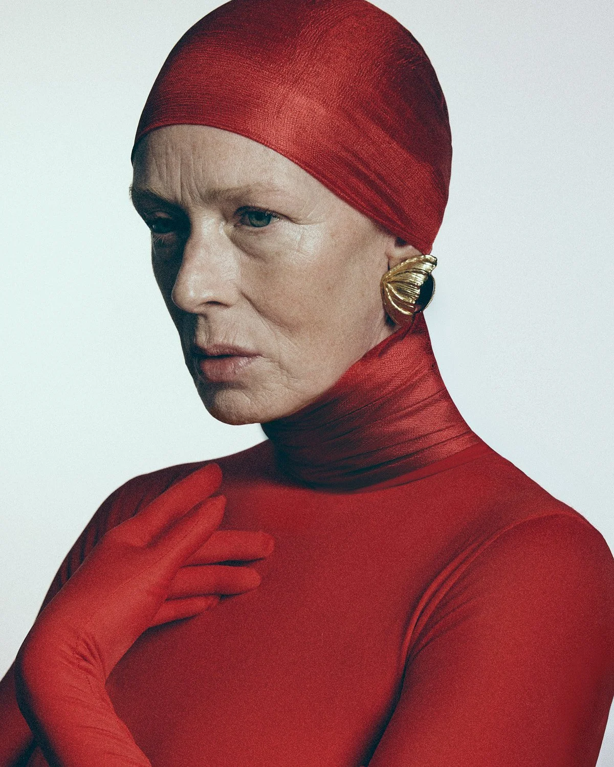 A woman is dressed in a red, form-fitting outfit and matching headscarf. She has a serious expression, with light makeup and prominent earrings. Her hand, covered with a red glove, is gently touching her chest.