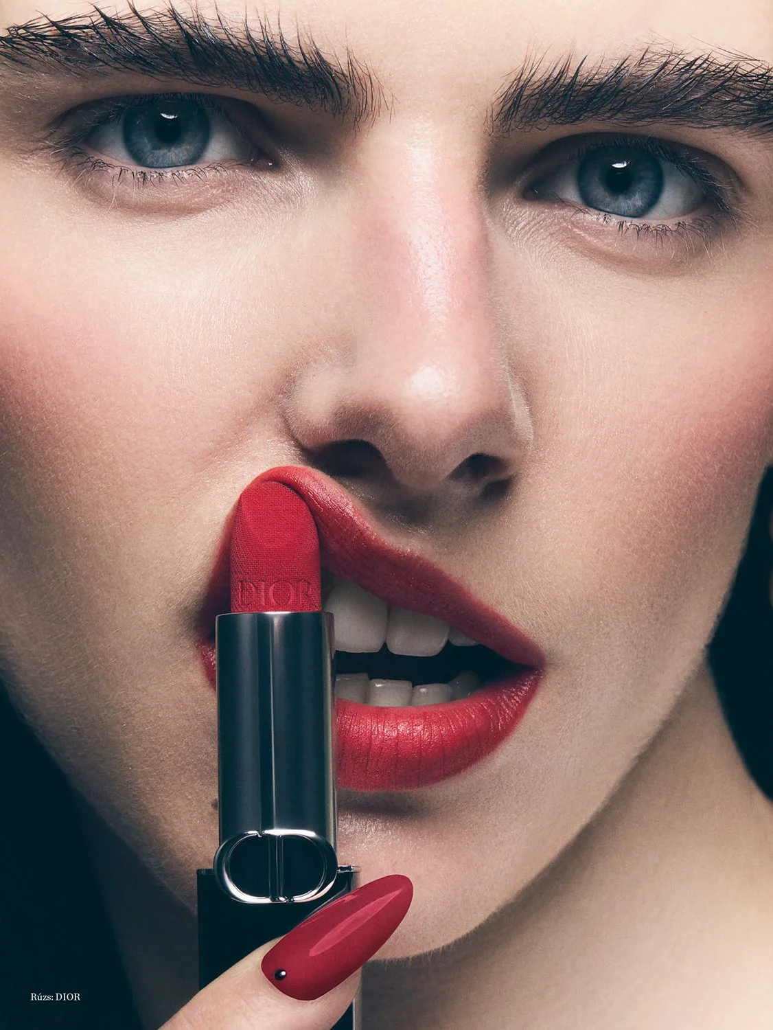 Close-up of a woman applying red lipstick with a Dior lipstick tube. She has blue eyes, well-defined eyebrows, and red lipstick on her lips.