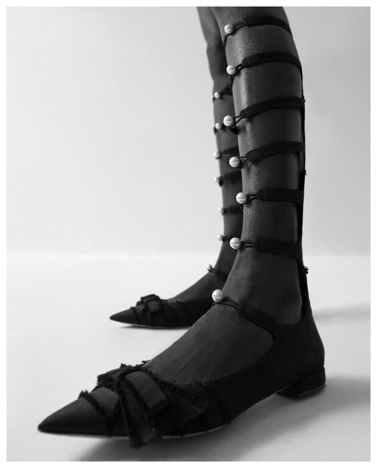 Black lace-up high heel boots with pointed toes, featuring elastic straps with pearl details running up the sides, worn with sheer black tights.