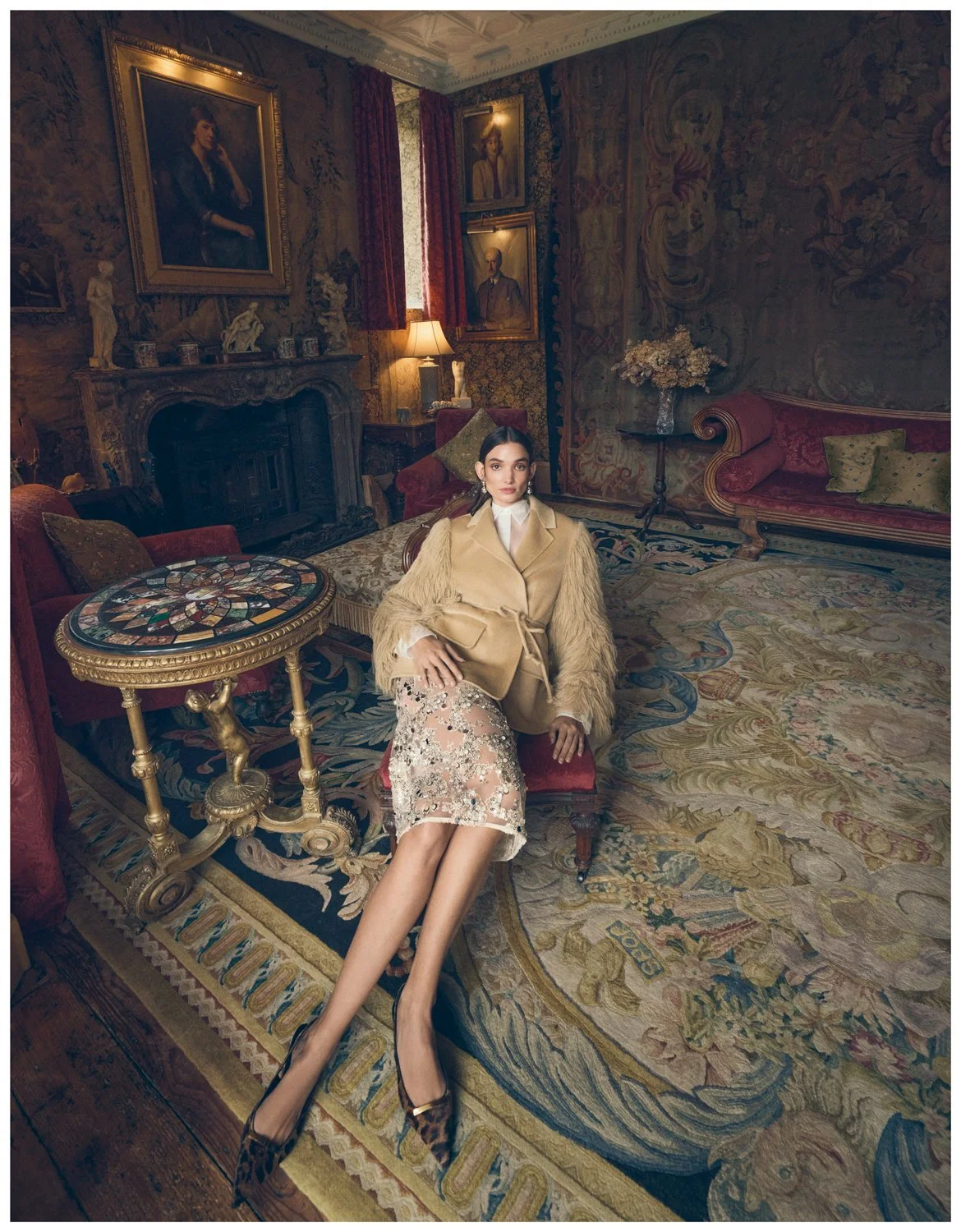 A woman with dark hair and wearing elegant clothing sits on a red velvet sofa in a richly decorated, historic room with paintings, a fireplace, and floral decor.
