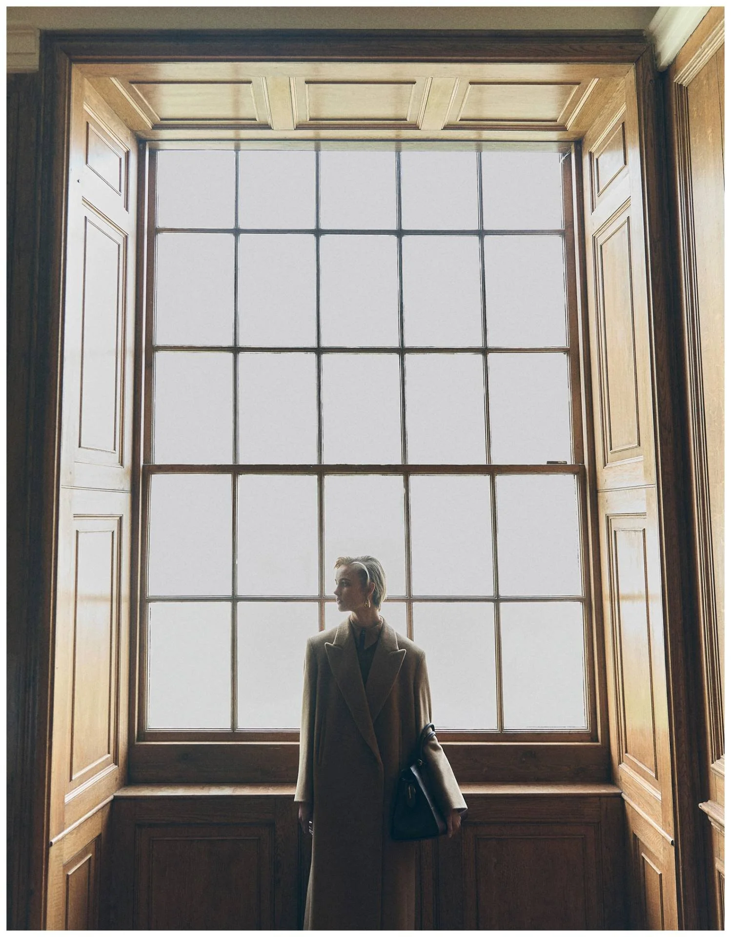 A woman standing in front of a large wooden-framed window, dressed in a beige coat, holding a black purse, with a soft indoor light illuminating her silhouette.
