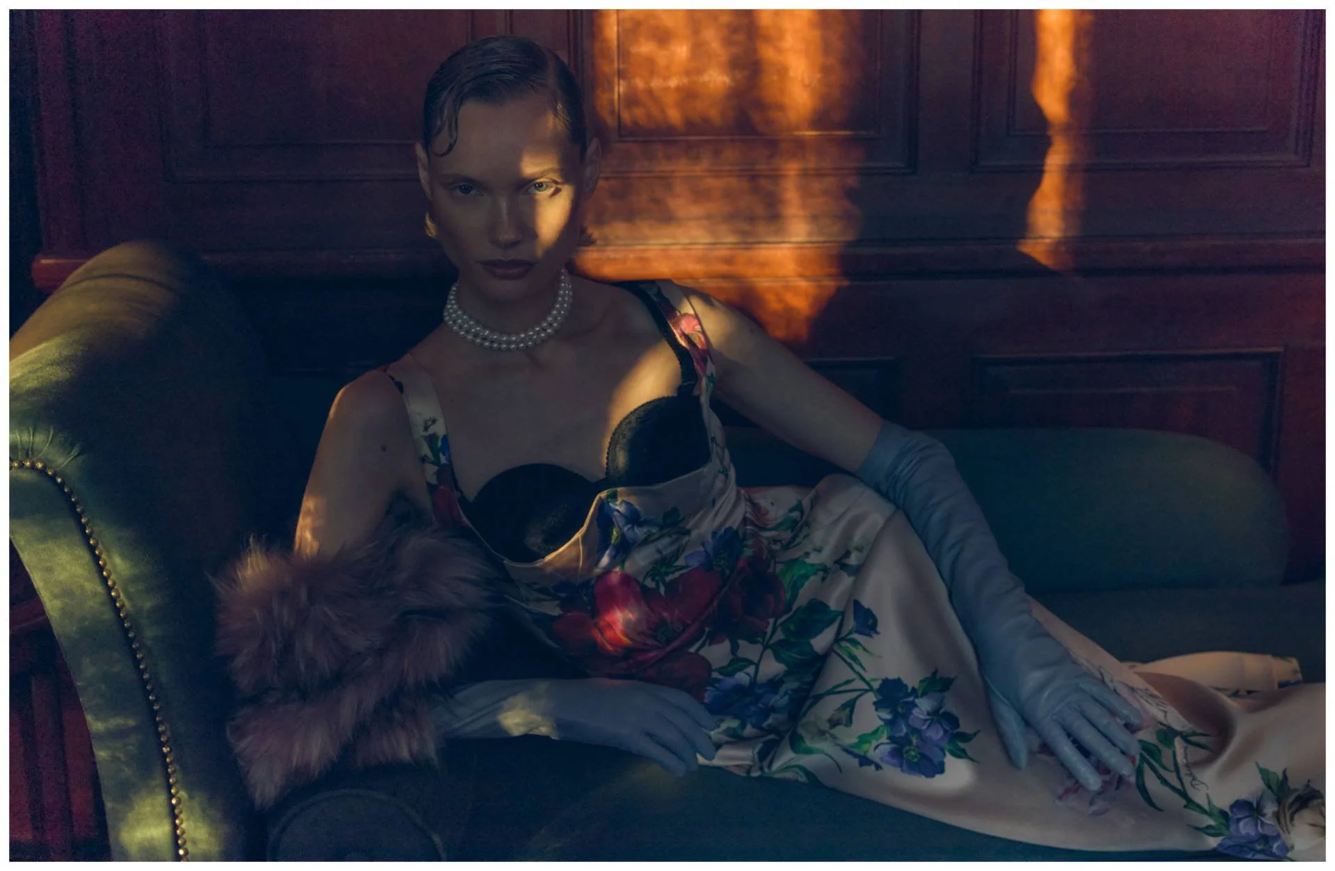 A woman in a floral dress and pearl necklace reclining on a vintage green study with wood-paneled walls, wearing long satin gloves, with a soft fox fur on her arm, illuminated by a dramatic light.