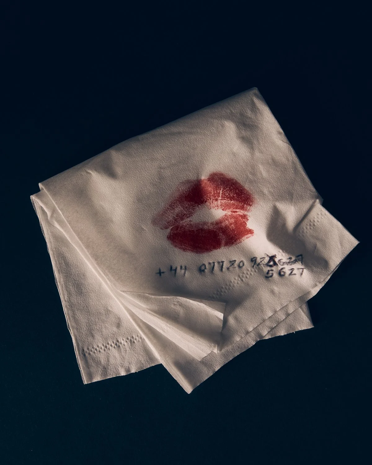 A white tissue with red lipstick kiss mark and handwritten phone number on a dark background.