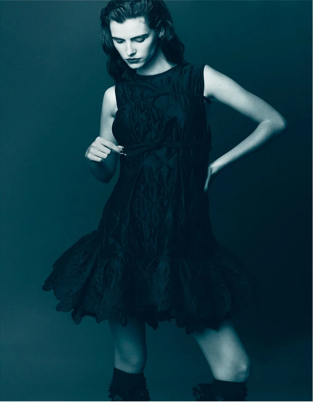 A woman with dark wavy hair wearing a sleeveless black dress with intricate patterns and ruffles at the hem, standing against a plain, dark background, looking down with one hand on her waist and the other holding the dress.