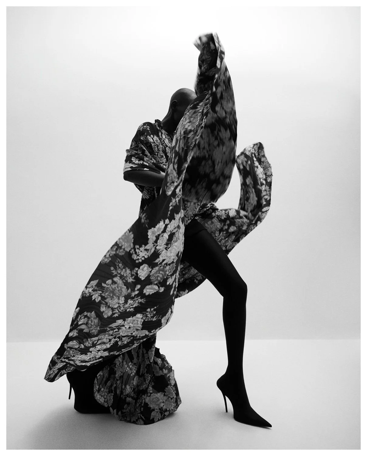 A black-and-white photo of a person with a shaved head posing dramatically, wearing high heels, with a flowing floral-patterned fabric wrapped around and lifted into the air.