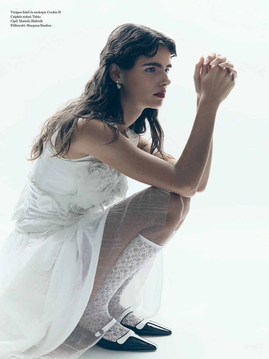 A woman dressed in white clothing with lace stockings and black shoes, squatting against a plain white background. She has dark, wavy hair and is looking to the side with hands clasped.
