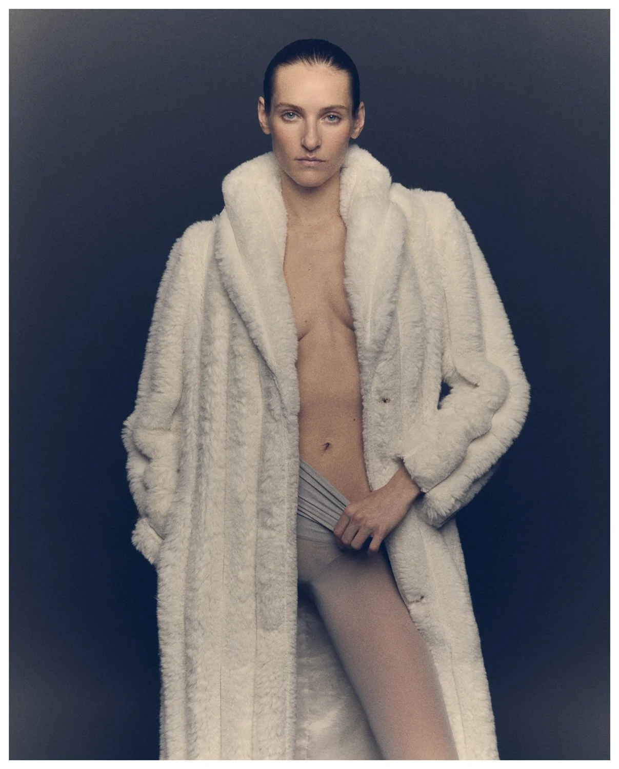 A woman with slicked-back hair wearing a large, white, fluffy coat with no visible shirt underneath, standing against a dark background.
