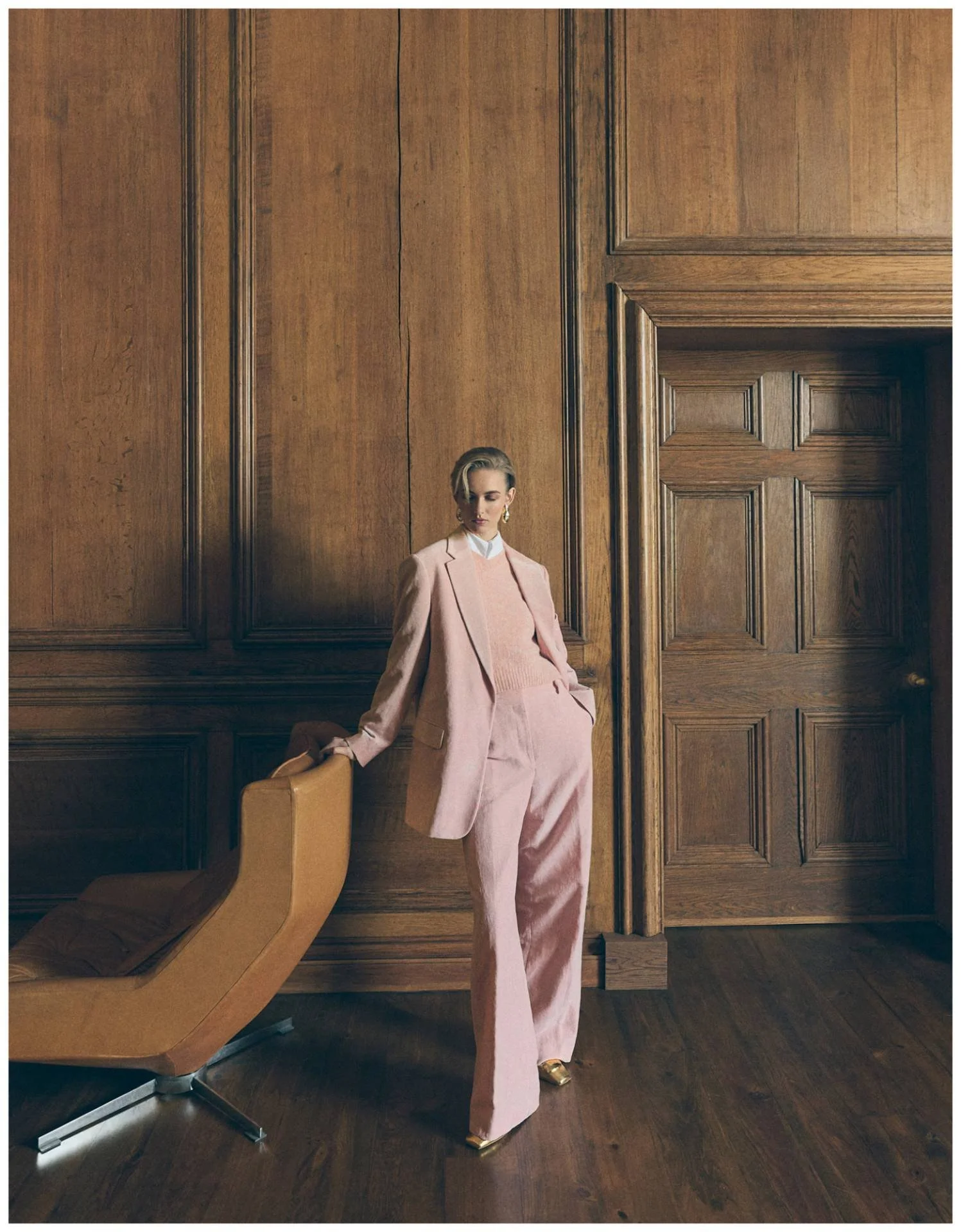 A woman in a pink suit and gold shoes standing in a wood-paneled room, leaning on a tan chair.