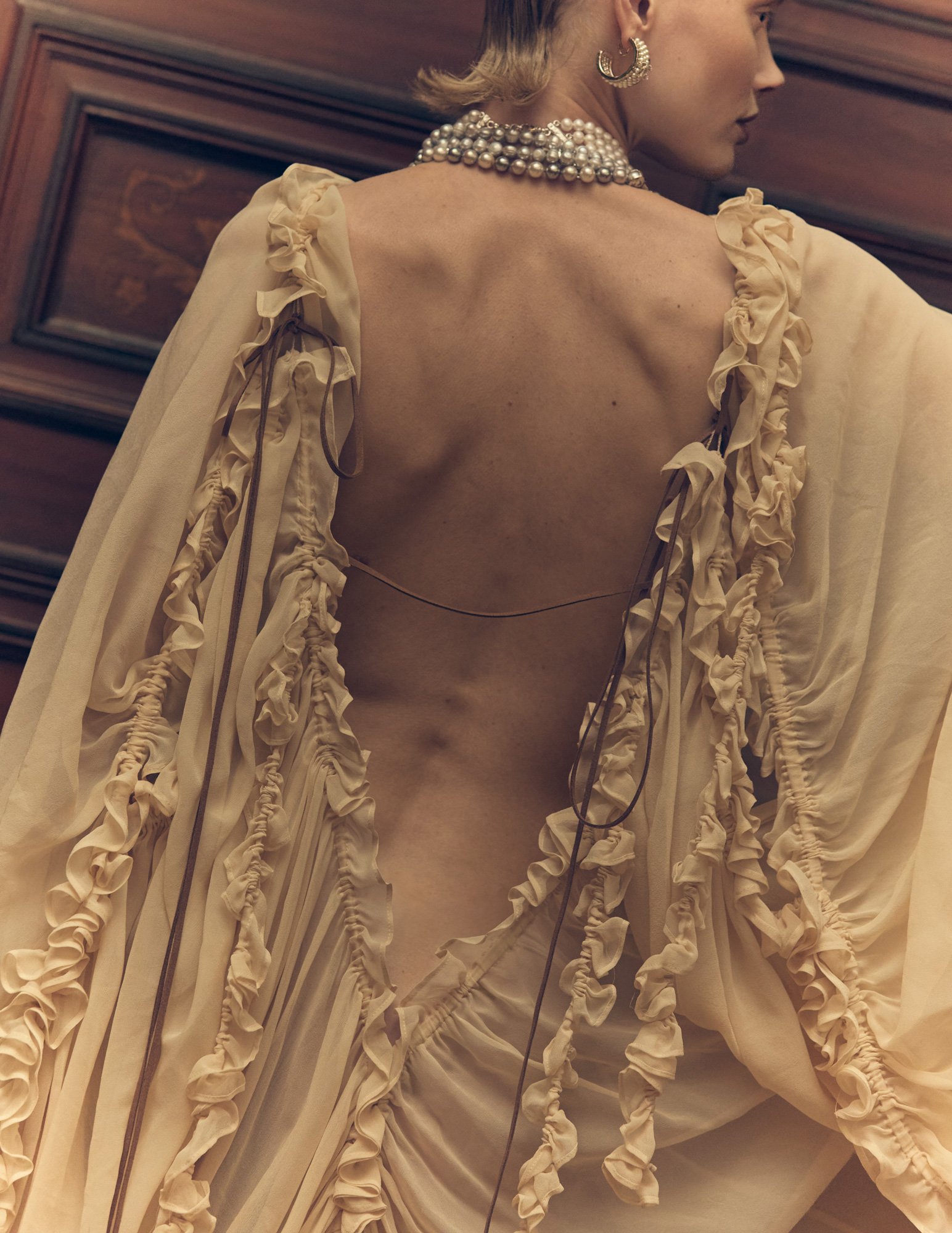 A woman dressed in an elegant, beige gown with ruffled details, wearing large pearl earrings and layered pearl necklaces, captured from behind in an indoor setting with wood-paneled walls.