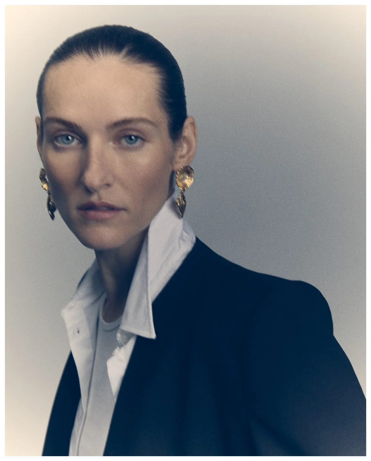 Close-up portrait of a woman with slicked-back hair, wearing large gold earrings, a white shirt with the collar visible, and a dark blazer, against a plain background.