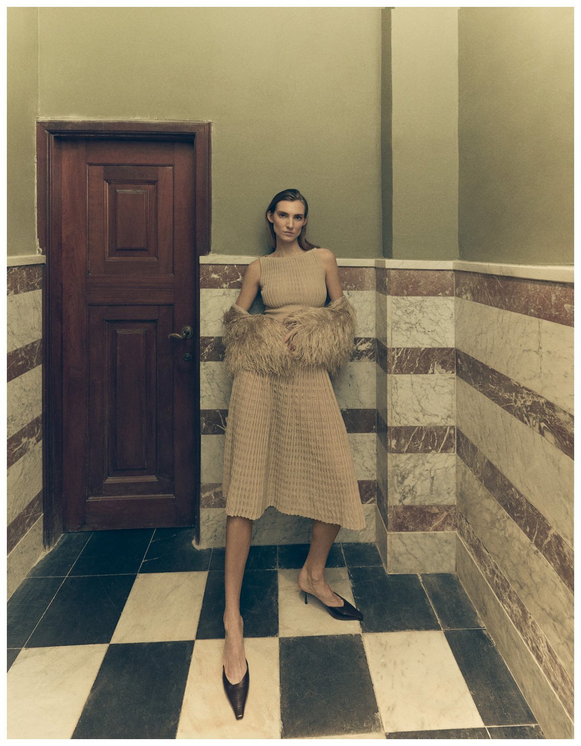 A woman standing in a corner of a room with marble walls and a checkered floor, wearing a beige knit dress with fur sleeves and high heels.
