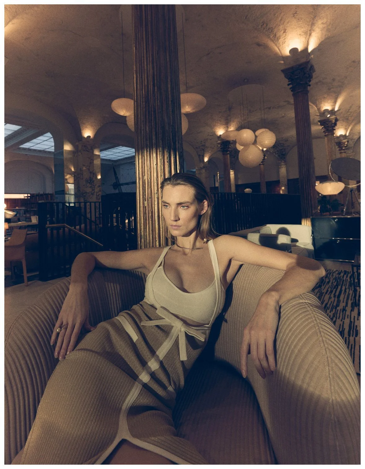 A woman in a beige tank top and striped wide-leg pants sitting on a beige upholstered sofa in a dimly lit, elegant interior with tall columns and hanging pendant lights.