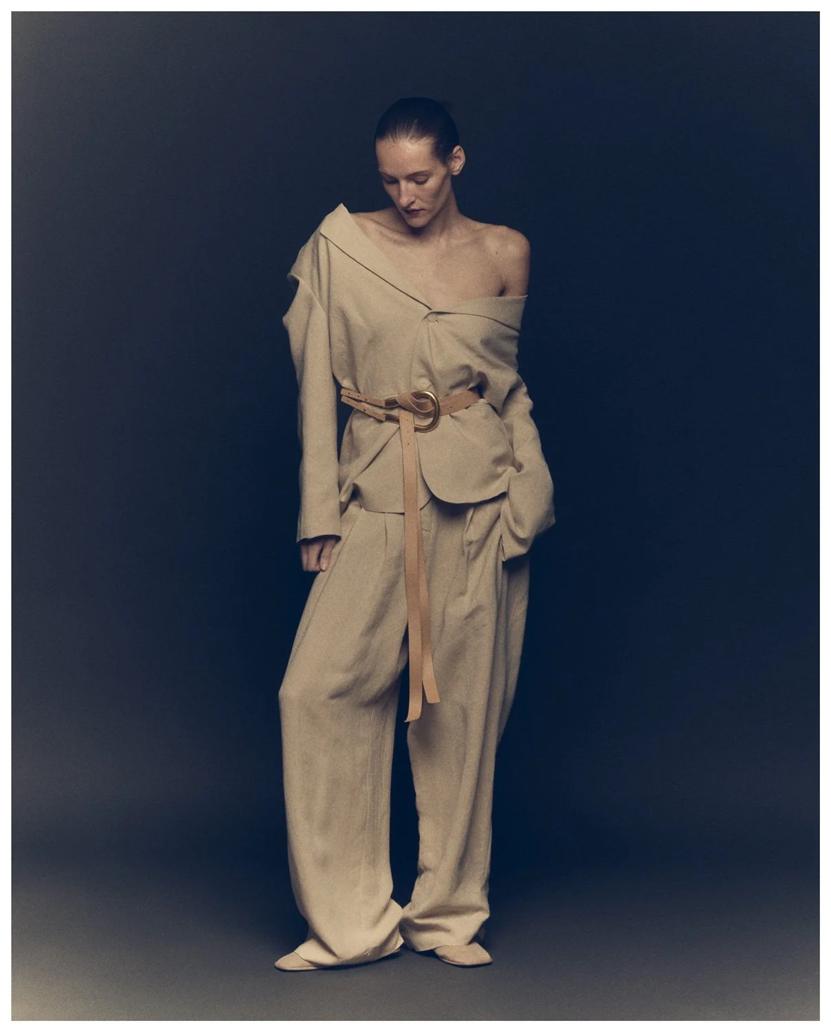 A woman with slicked-back hair wearing a beige oversized blazer with one shoulder exposed, matching wide-leg pants, and a beige belt with a large circular buckle, standing against a dark background.
