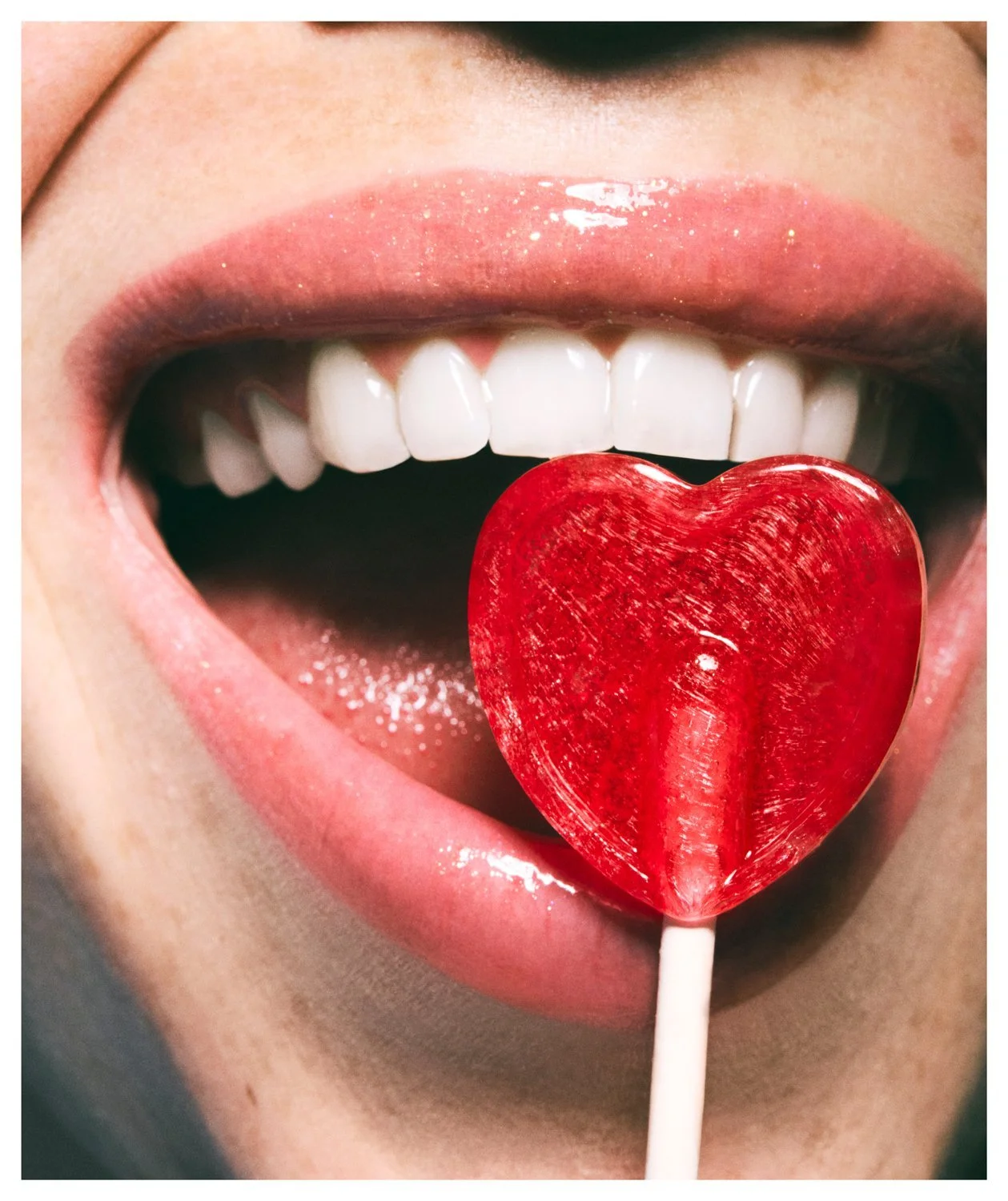 Close-up of a person's open mouth with red lip gloss and white teeth, holding a red heart-shaped lollipop on their tongue.