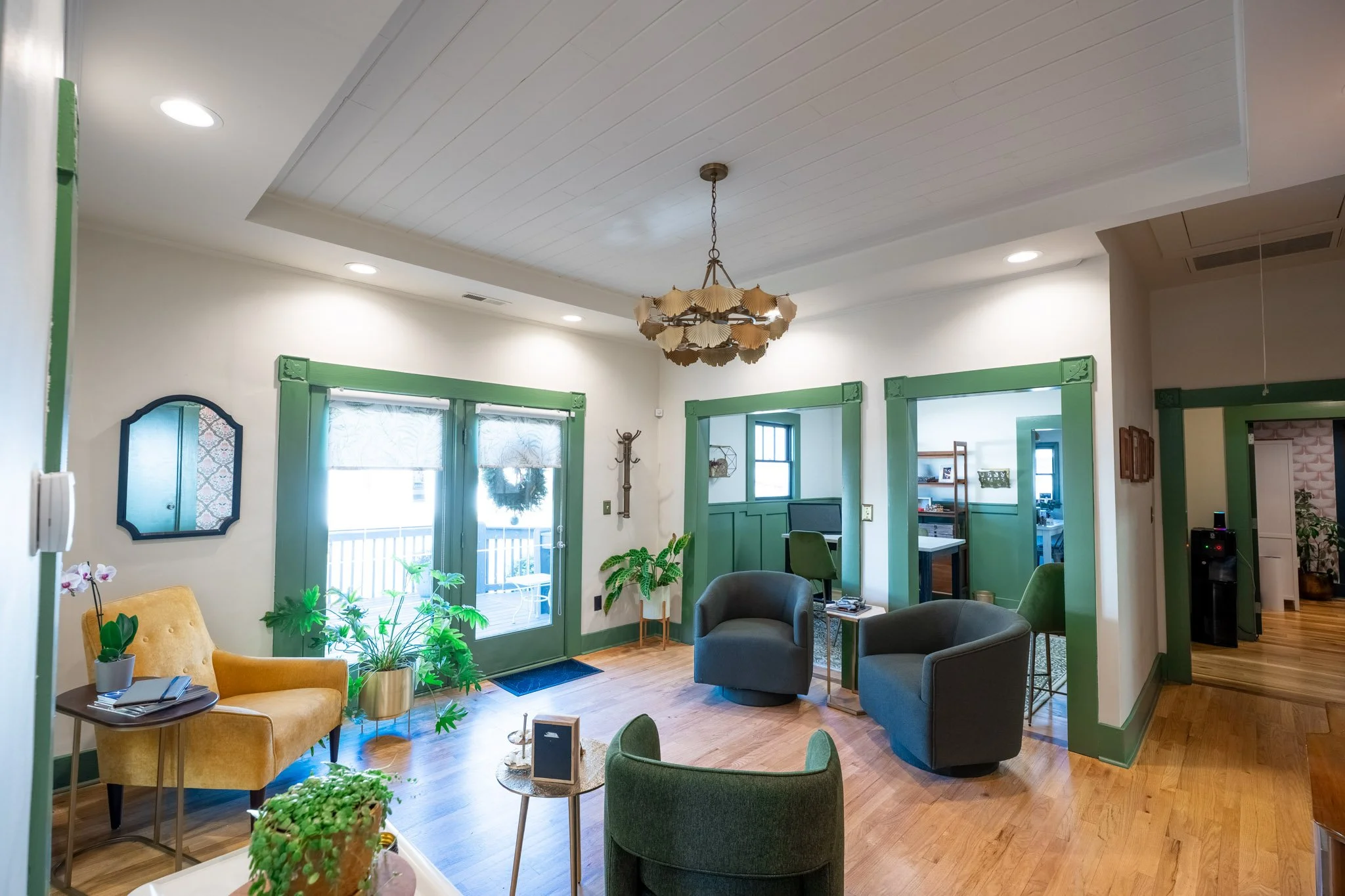 A bright living room with a white ceiling, green trim around windows and doors, and hardwood floors. There are various chairs including a yellow armchair, gray armchairs, and a green chair, along with plants, a coffee table, and decorative items. A c