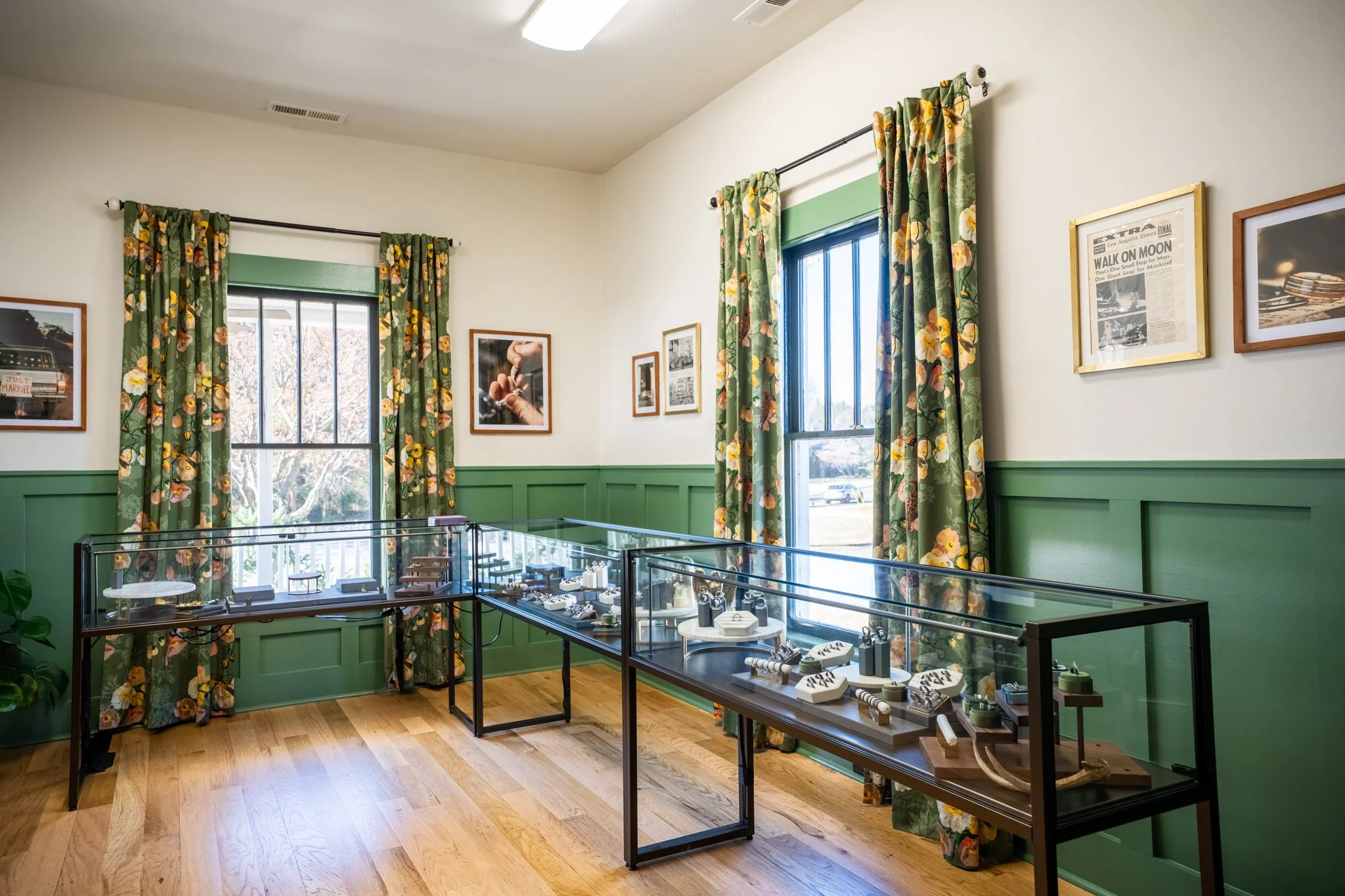 Room with green wainscoting, two windows with floral curtains, and various framed pictures on the wall. Glass display cases containing jewelry and small collectibles are in the foreground. Wooden floor and ceiling light fixture.