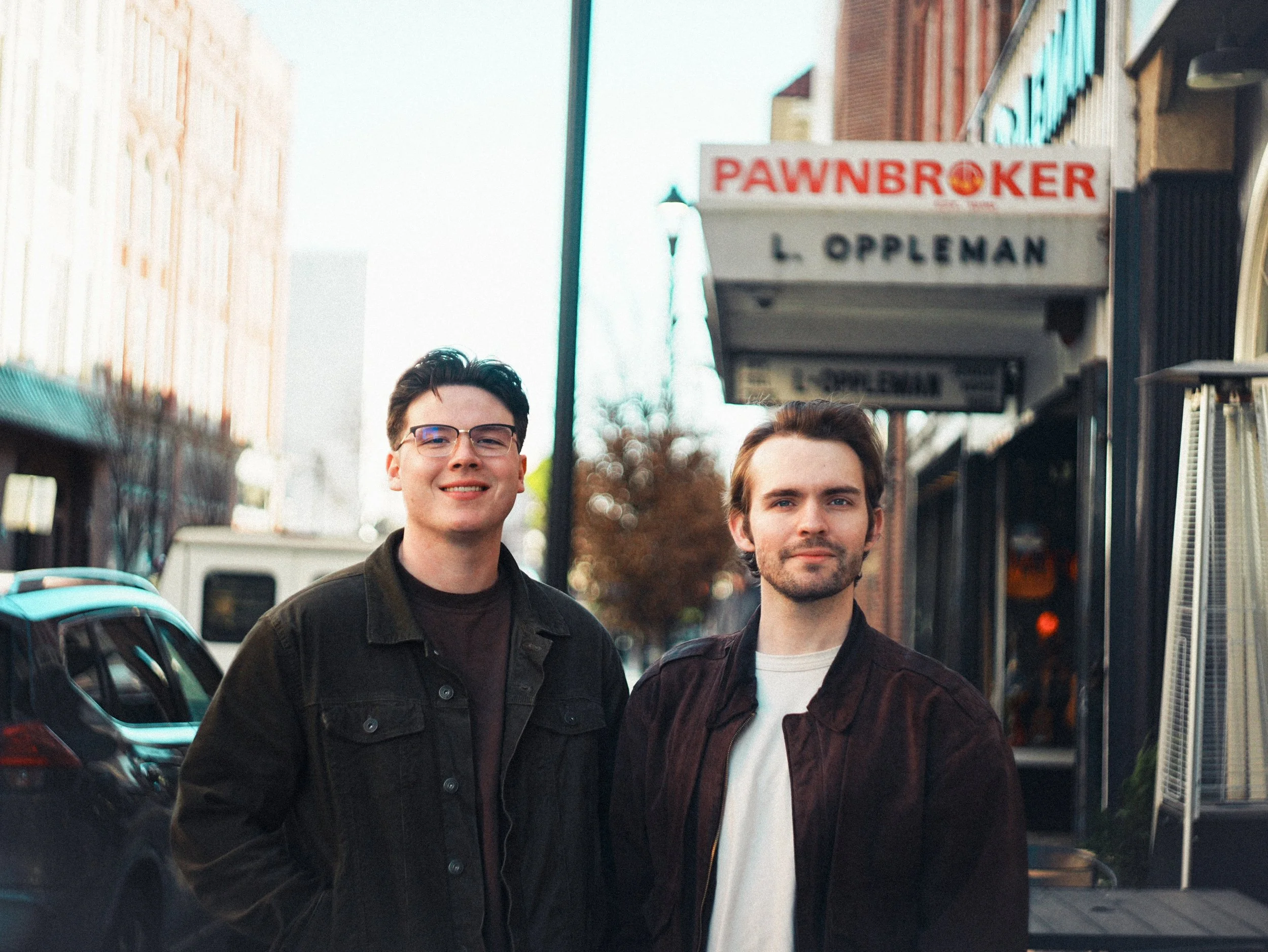 Two young men standing on a city sidewalk in front of parked cars and storefronts, with a "PAWNBROKER" sign and a taller building in the background.