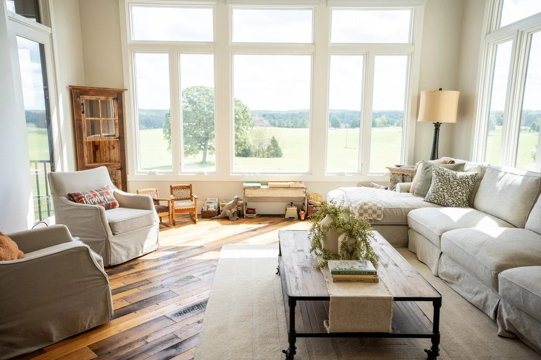 Bright living room with large windows overlooking a green landscape, beige sectional sofa, armchair, wooden coffee table, and various decorative items.