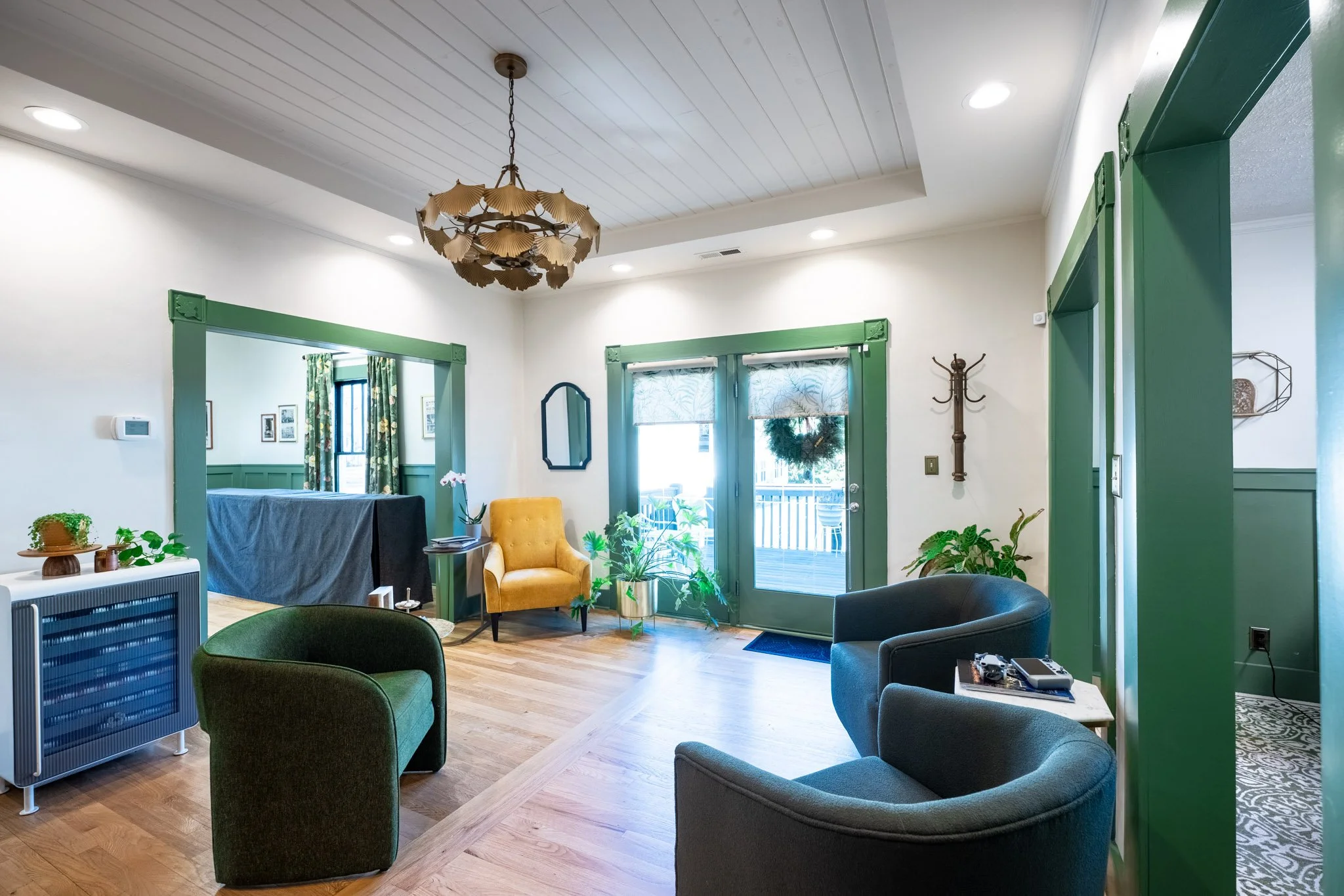 A cozy living room with green trim, hardwood flooring, and modern furniture including a yellow armchair and a gray sofa. There are multiple plants, a wooden coffee table, a small side table with electronic devices, and a large window with floral curt
