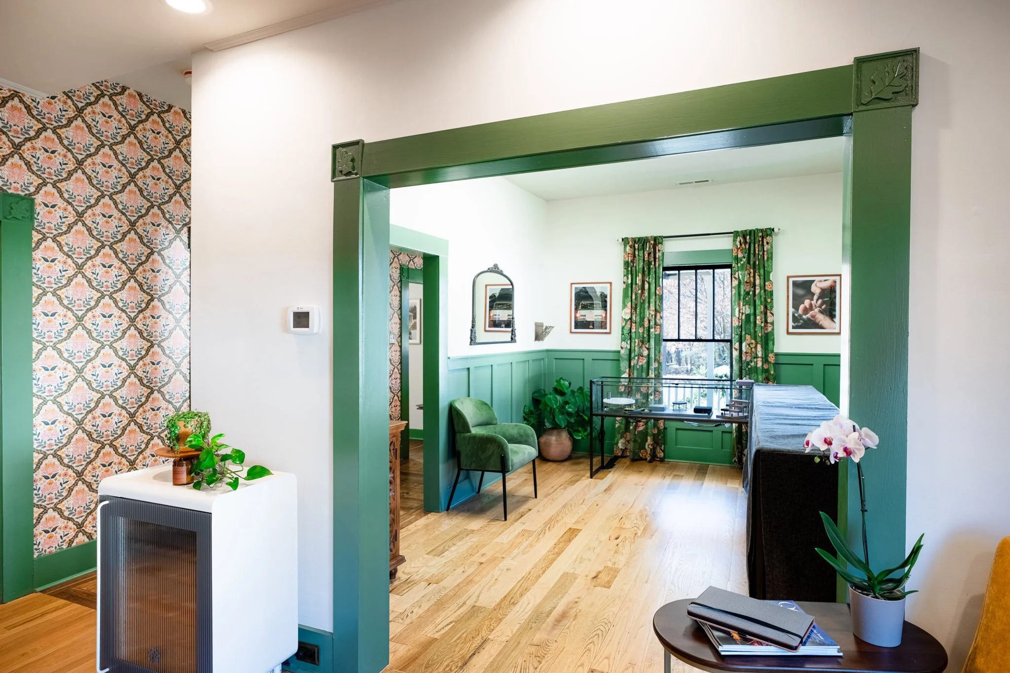 A cozy living room with green accents, floral curtains, and wooden flooring, visible through a doorway with decorative green trim.