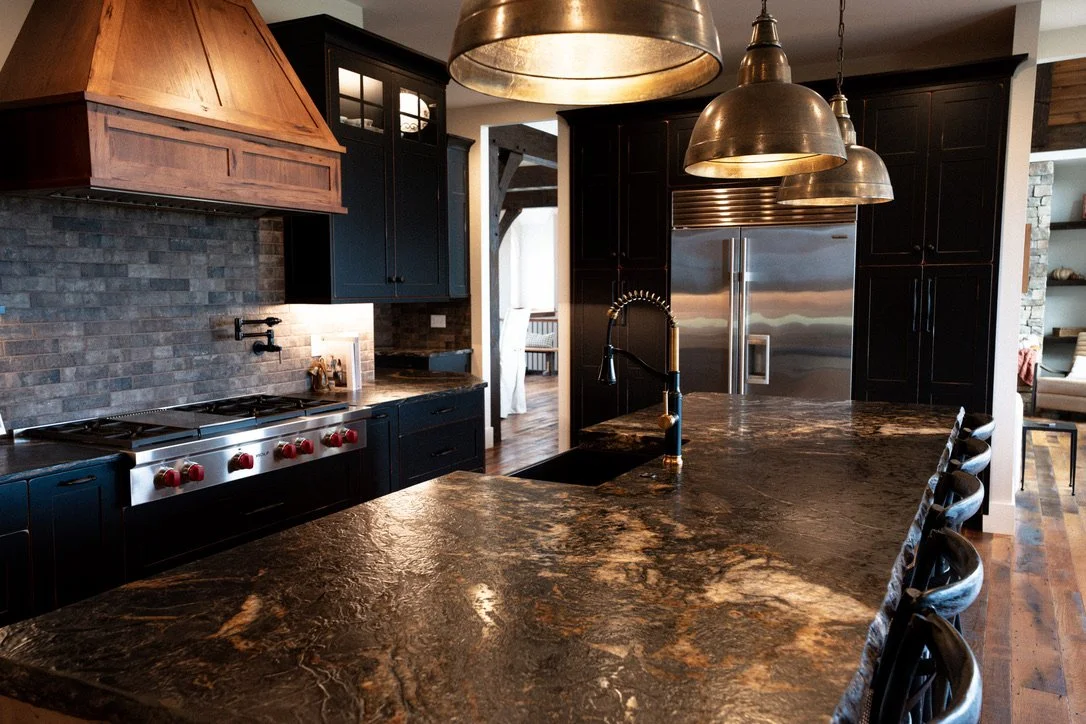 Modern kitchen with black cabinets, stainless steel refrigerator, stone countertops, and pendant lighting.