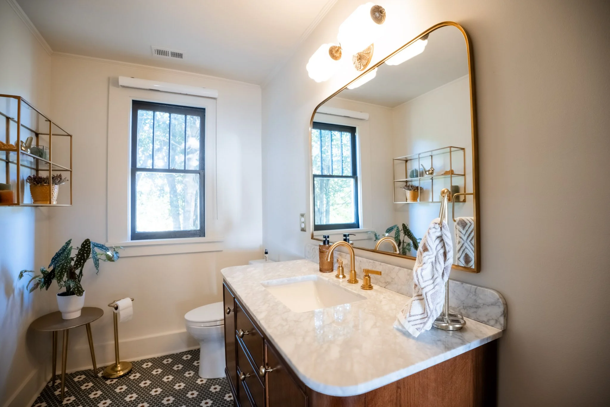 A modern bathroom with a marble countertop, gold fixtures, a large mirror, and window letting in natural light. Decor includes a plant, wall shelves, and bathroom accessories.